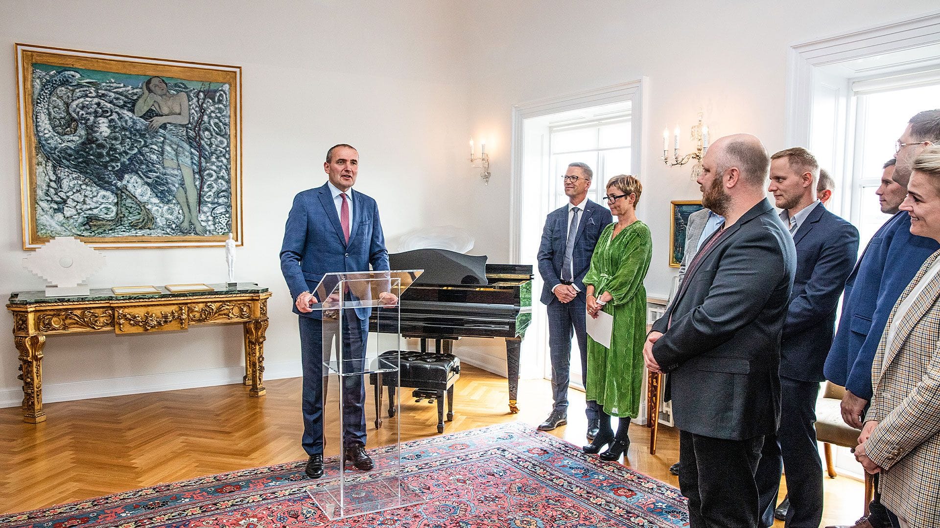 A man in blue suit speaking at a lectern in an elegant rom with artwork, a piano and attentive listener