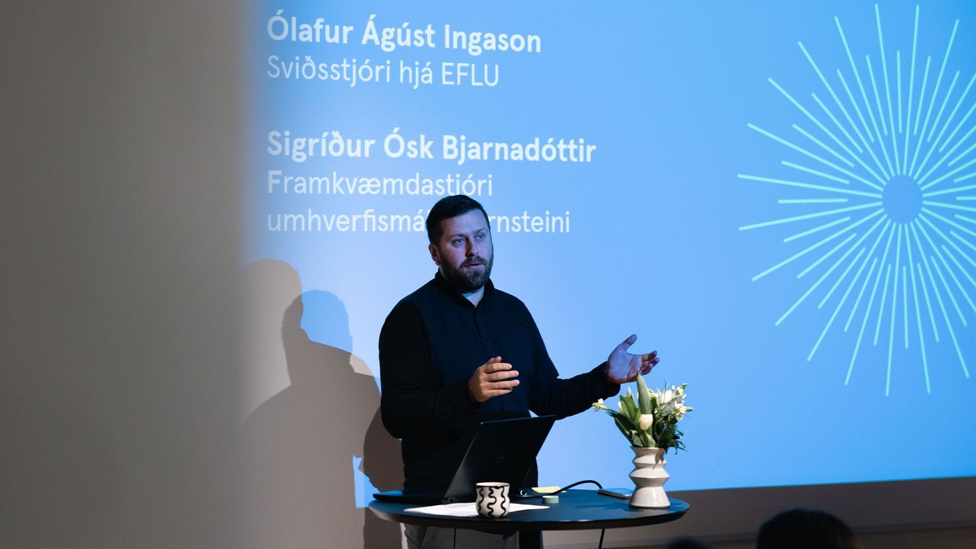 A man is presenting with a big screen in the background
