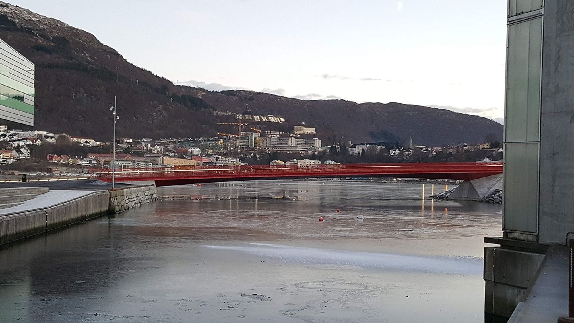 A red bridge against a backdrop of a calm body of water and hilly terrain