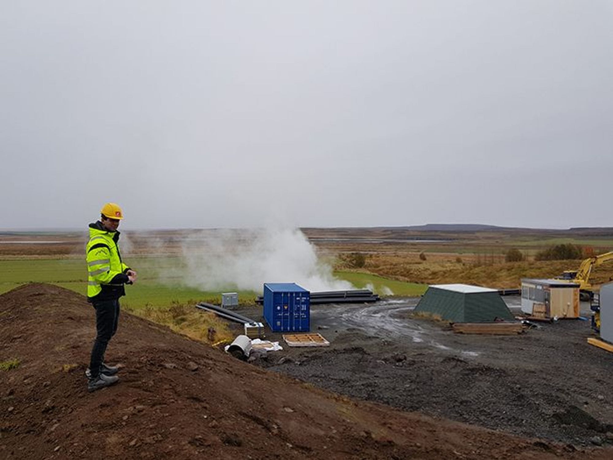 A person with high-visibility jacket standing on a geothermal site with steam vent 