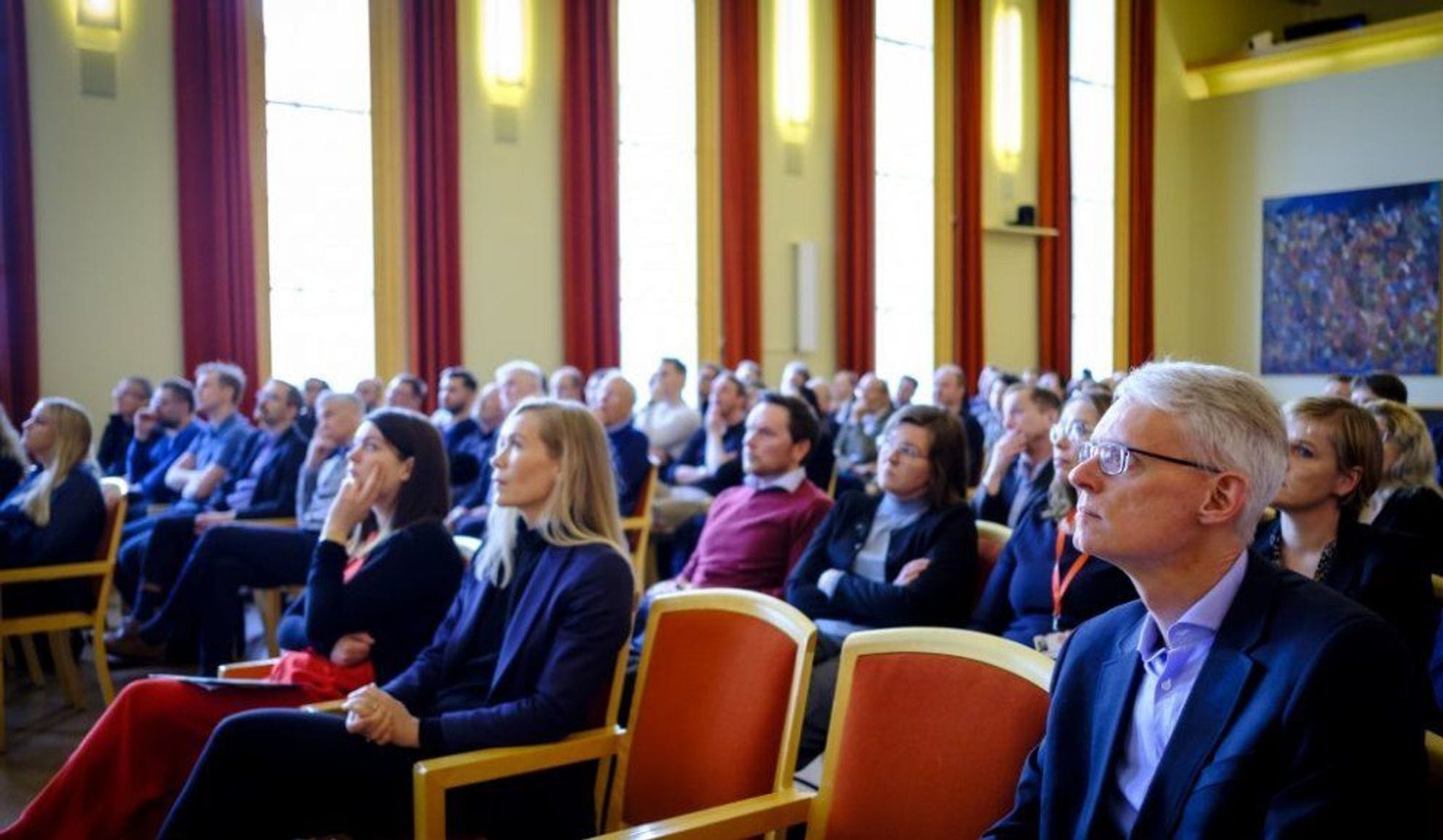 An audience of professionals looking straight and seated on red chairs 