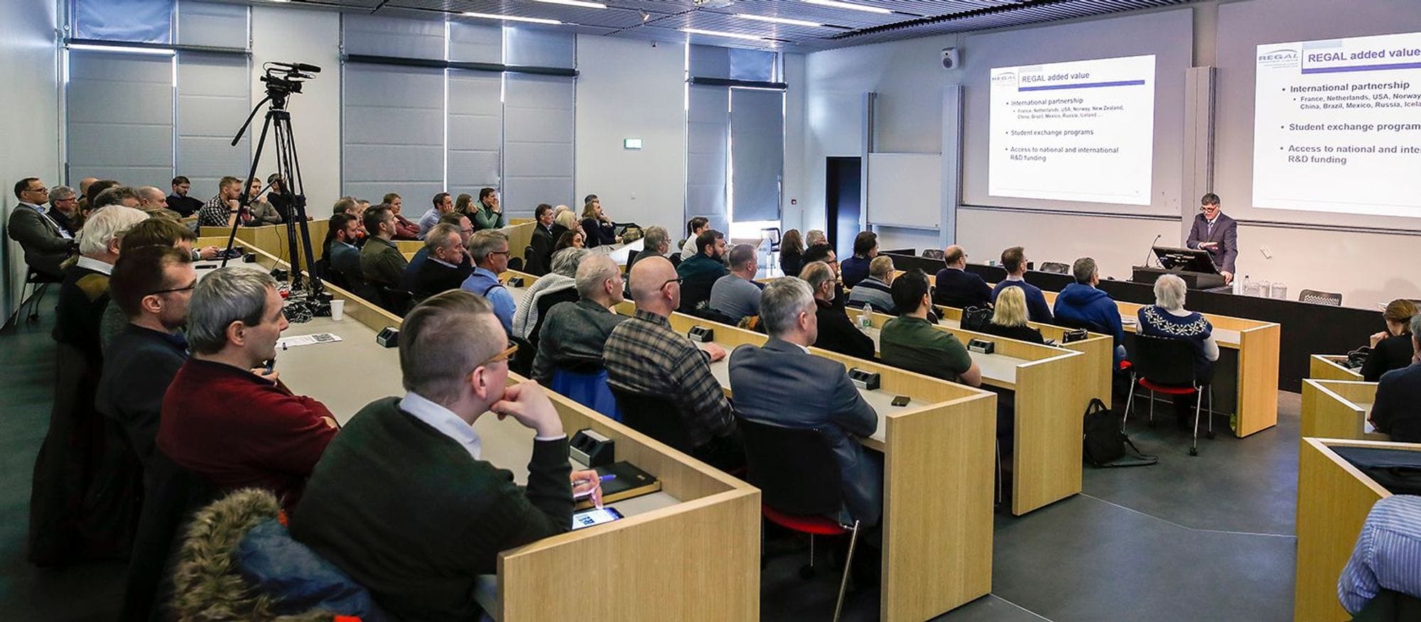 people seated on wooden desk, listening carefully to a man giving presentation 