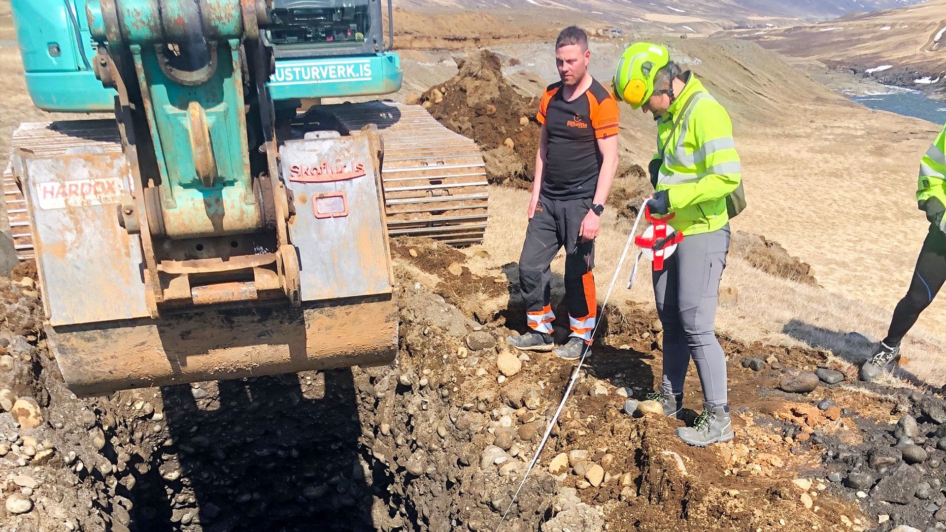 Two individuals, one in high visibility jacket and the other in an orange and black shirt, standing near an excavator