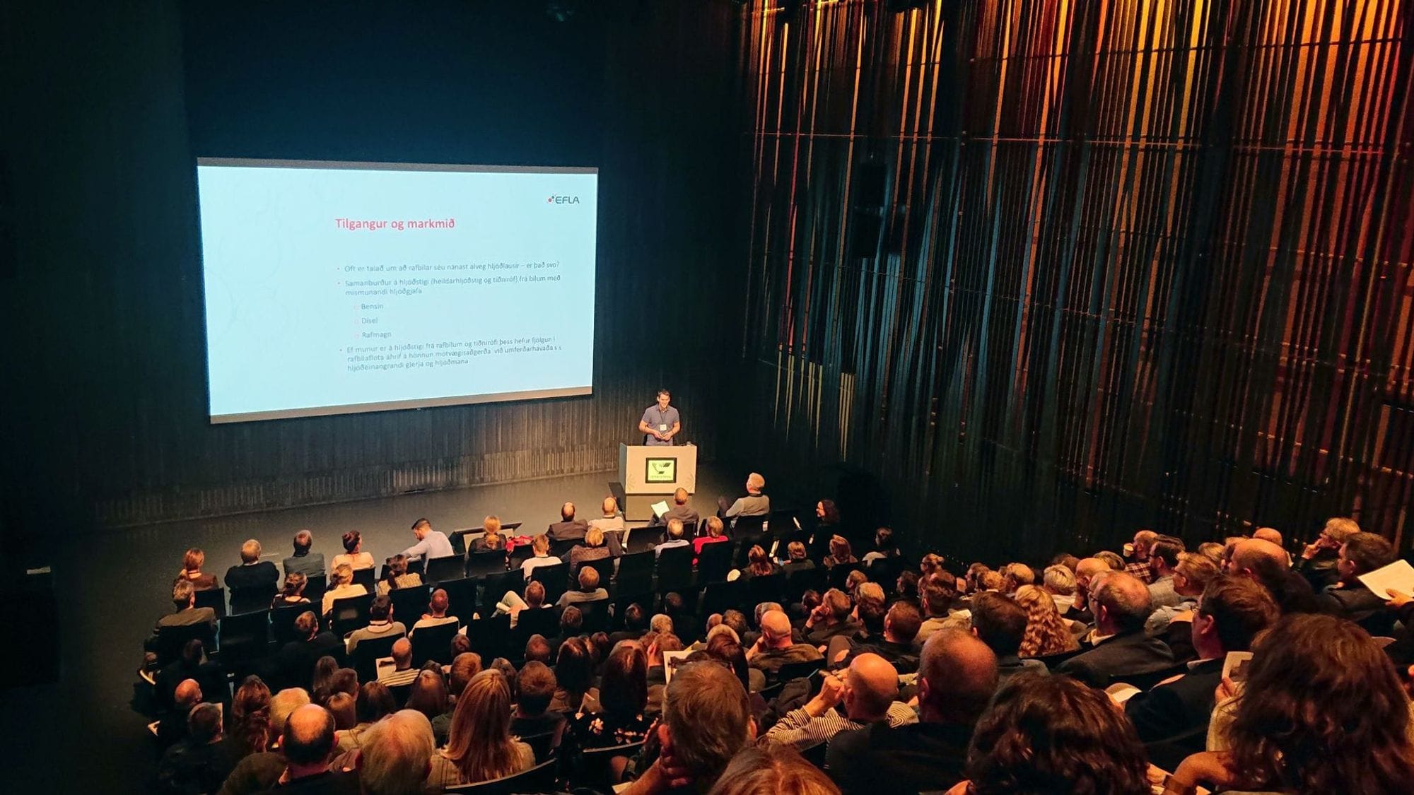 A large audience facing a speaker who is presenting in front of a large screen