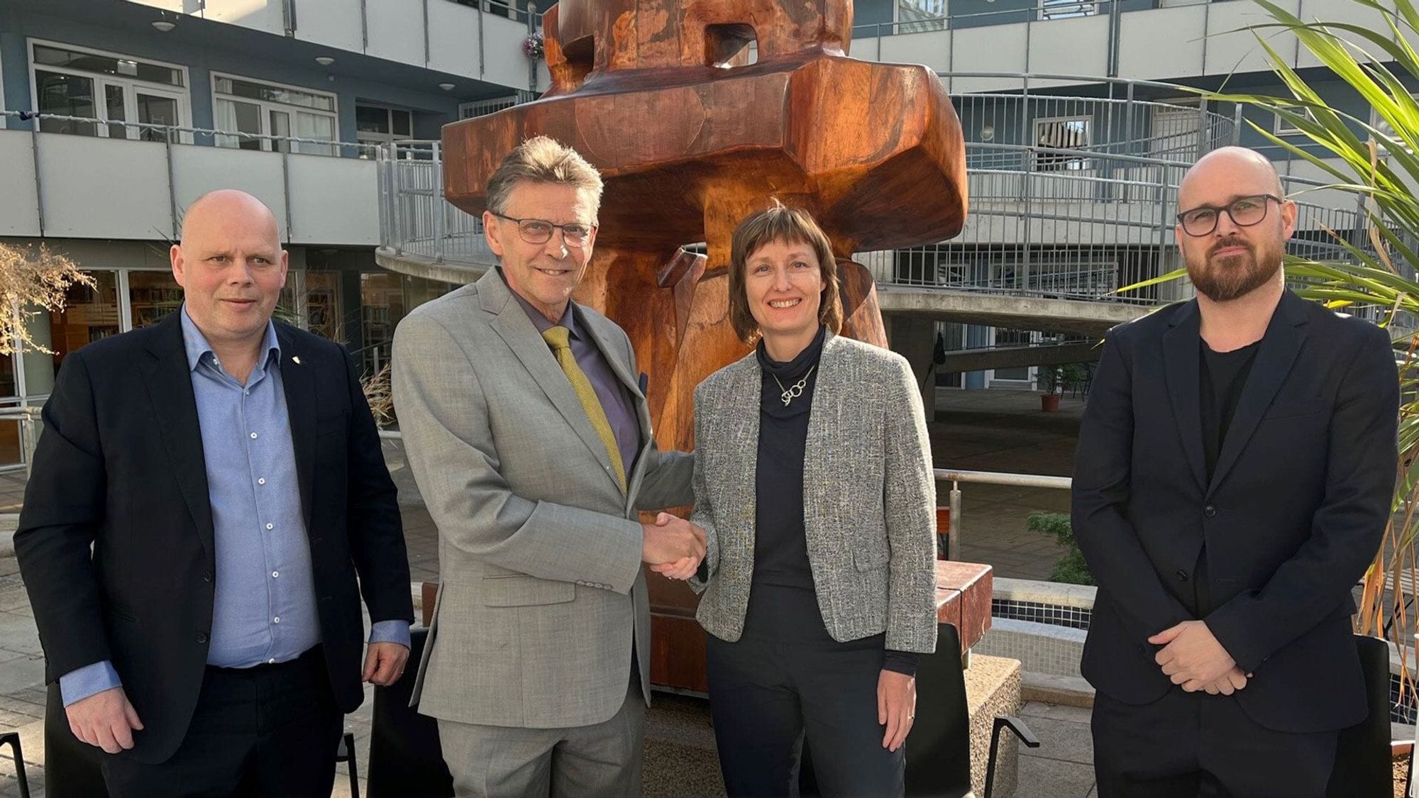The photo shows four individuals, all in professional attire, standing in front of a building with a large rusted metal sculpture