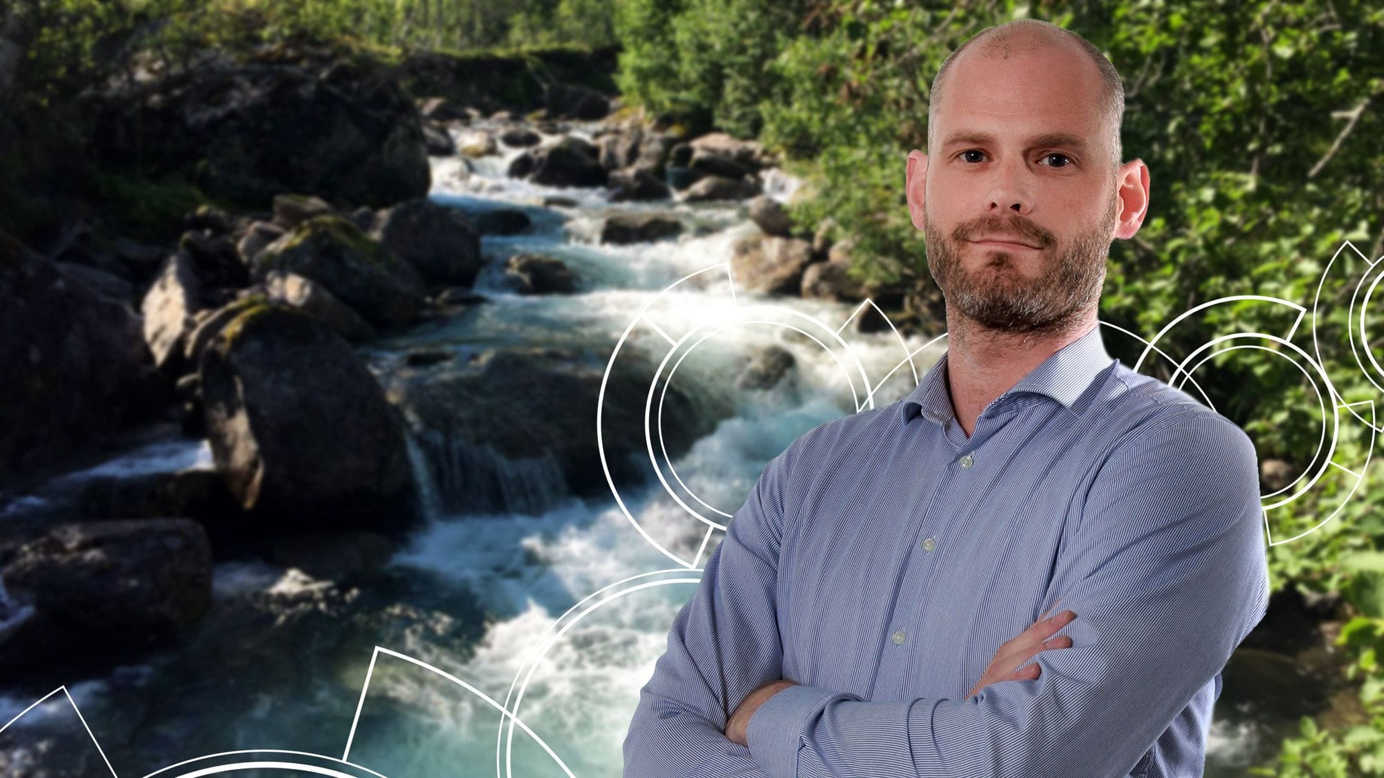 A man in blue shirt with his arm crossed in front of a blurred background featuring a rocky stream