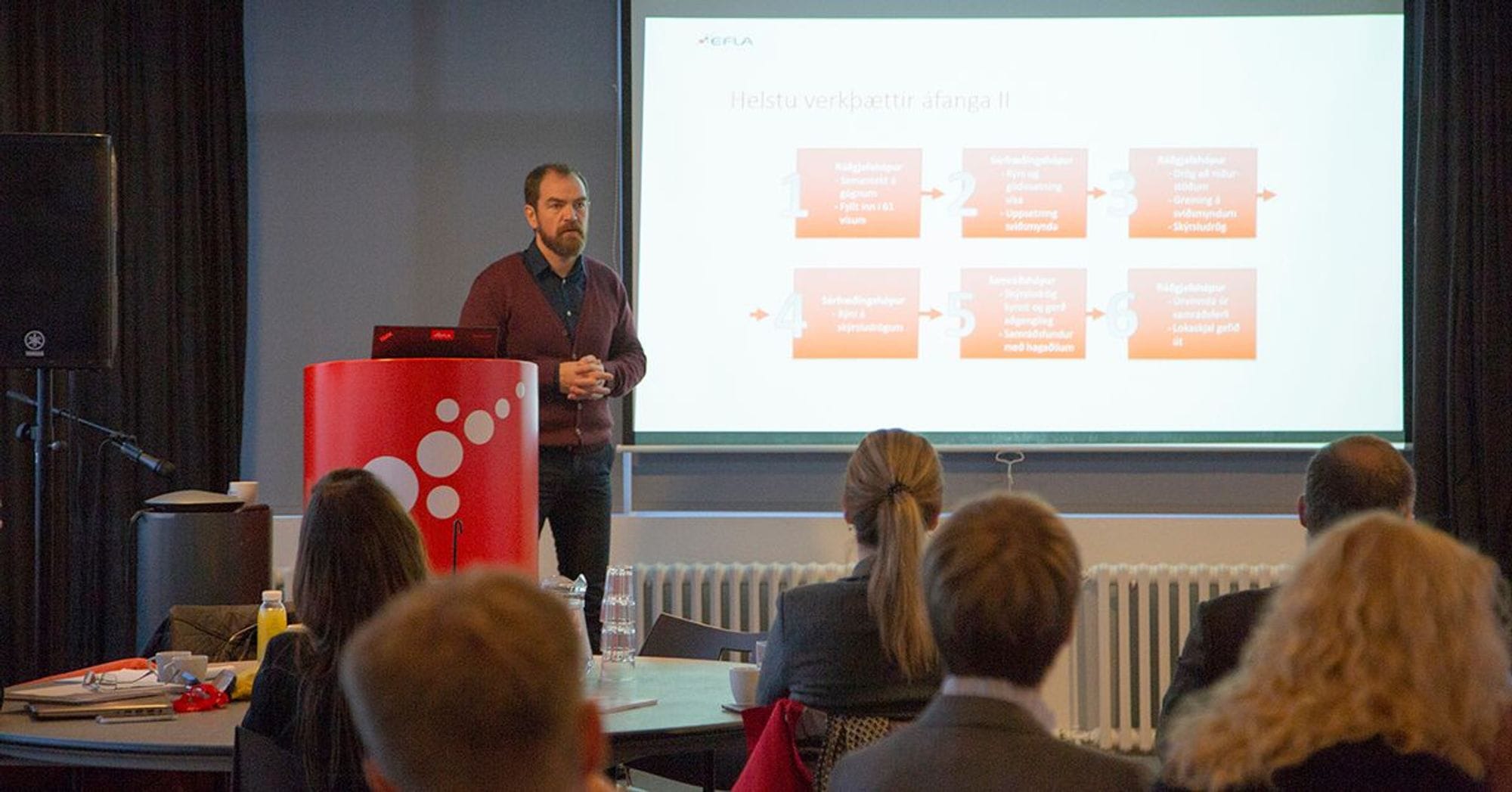 A man presenting to an audience, standing next to a screen