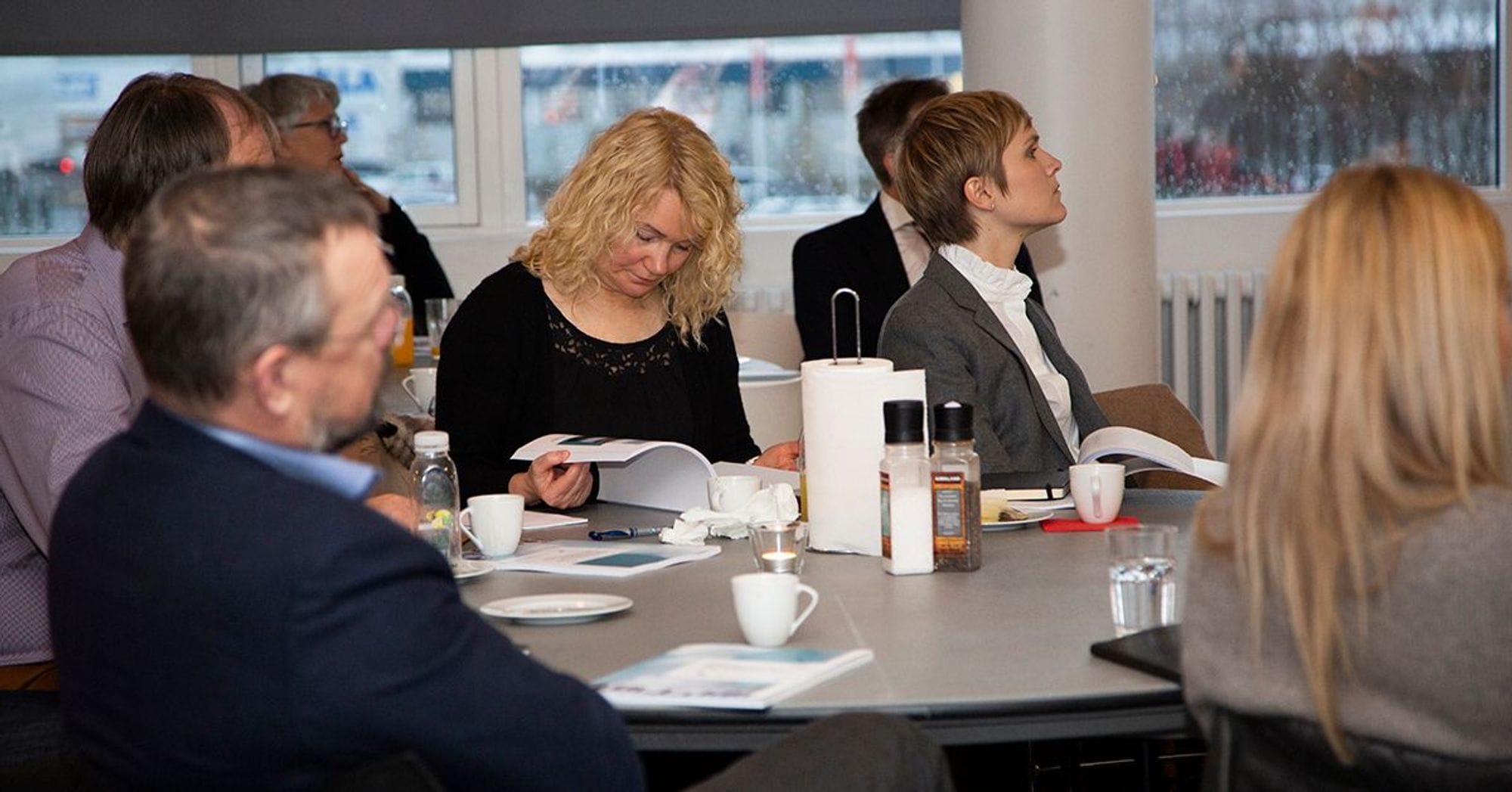 Individuals seated in a room attentively looking at the same direction and some reading  documents