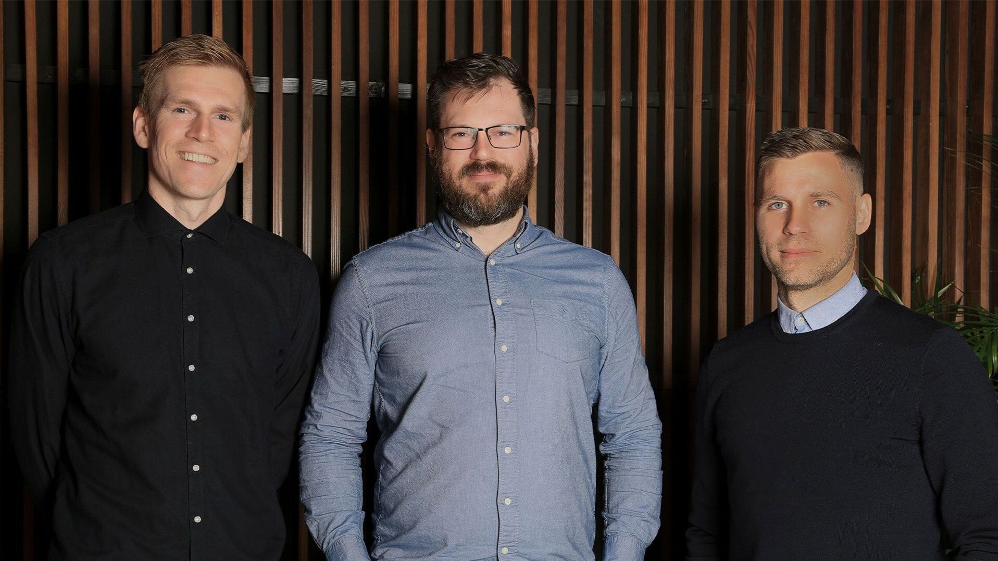 Three men standing together, smiling with a wooden slat wall in the background
