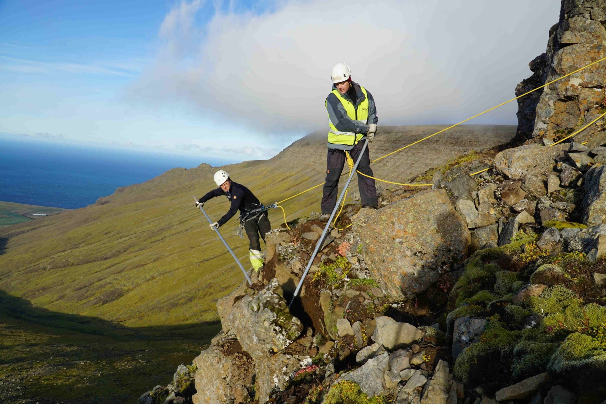Two individuals wearing high visibility jacket, working on a rocky hillside with rope