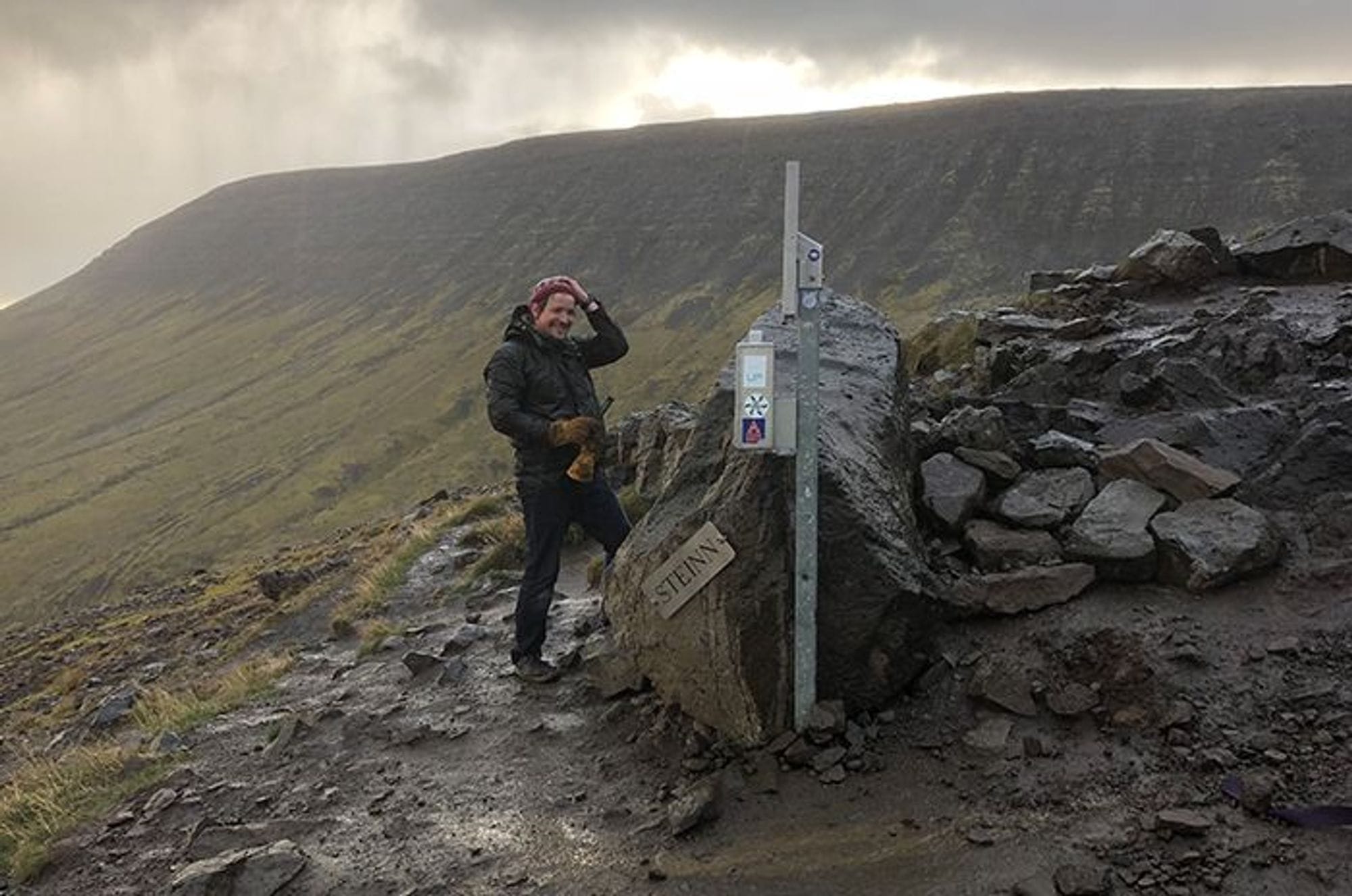 A man in outdoor clothing, smiling and standing besides a trail marker