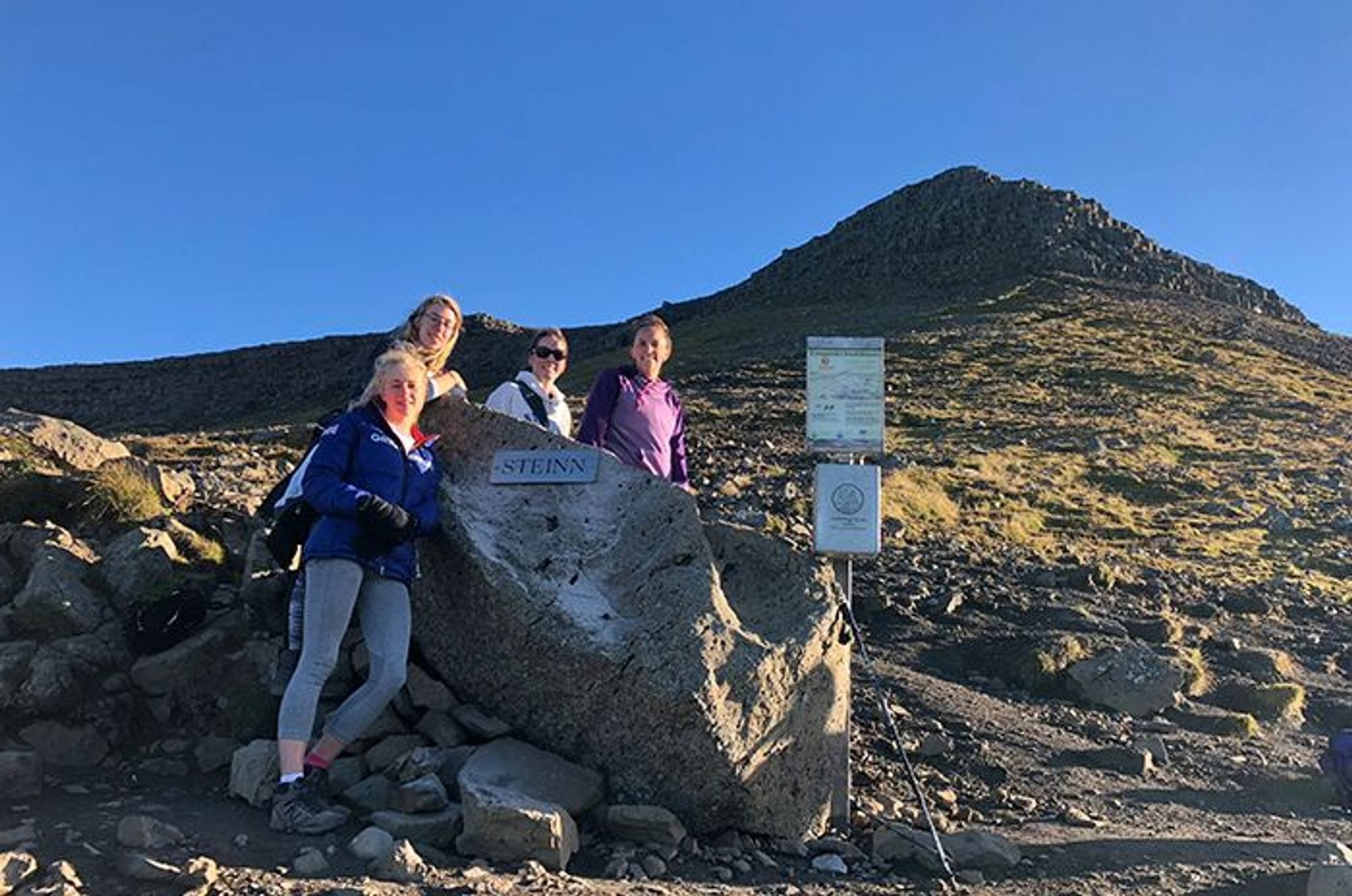Four women standing next to a large rock with a sign labeled "STEINN"