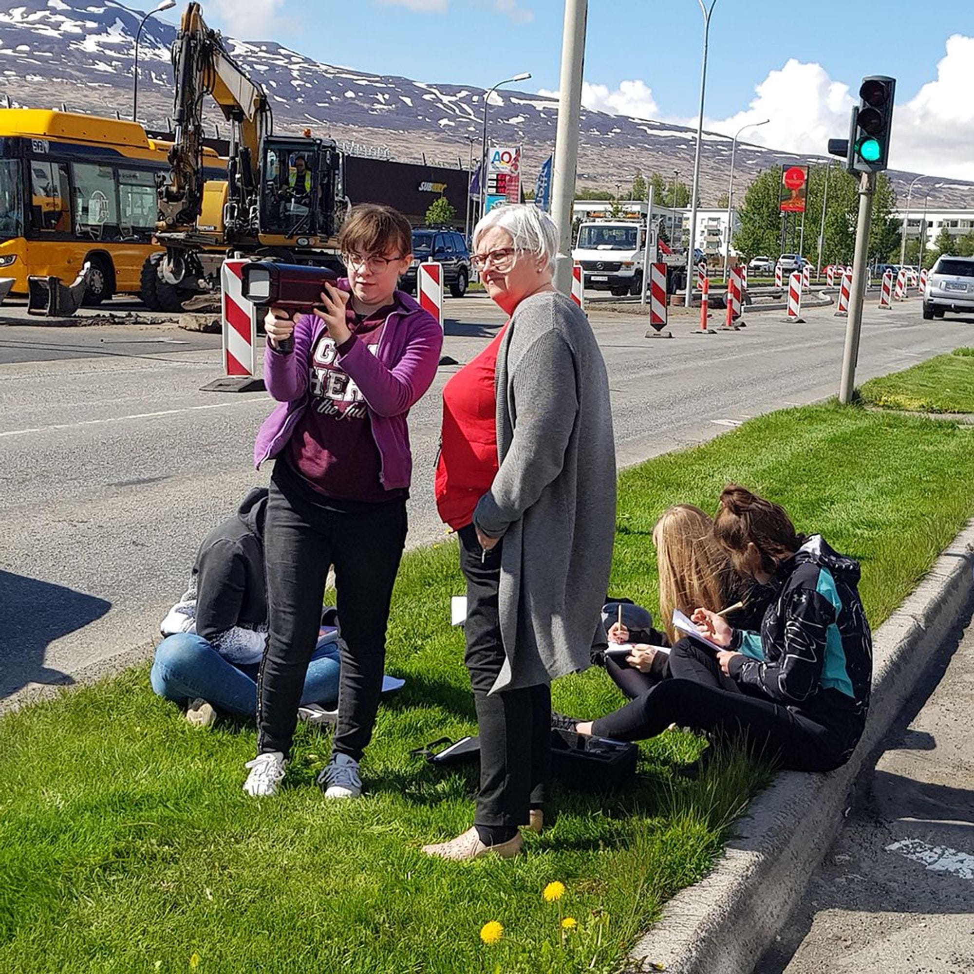 Two women examining car speed reading machine and other people sitting at the background, writing on notebooks