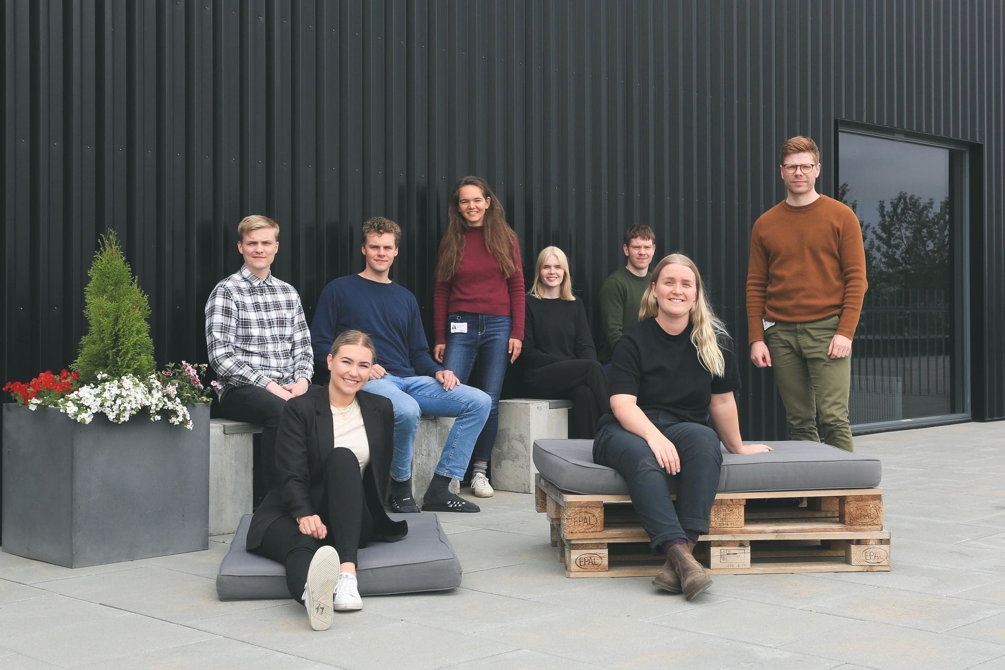Several individuals posing for a photo outdoor, with some seated on a pallet benched and others standing