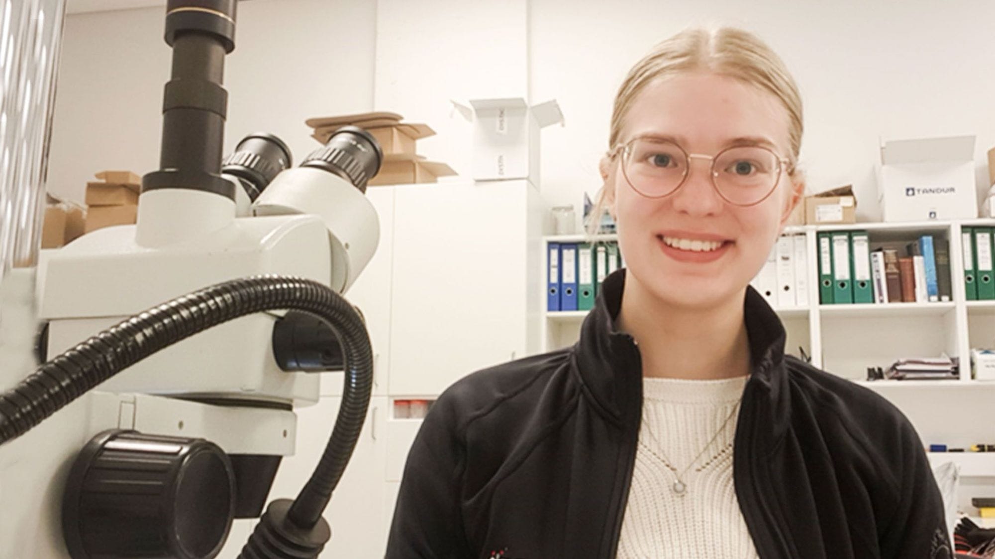 A smiling people with glasses positioned in a laboratory setting with a microscope and bookshelves in the background