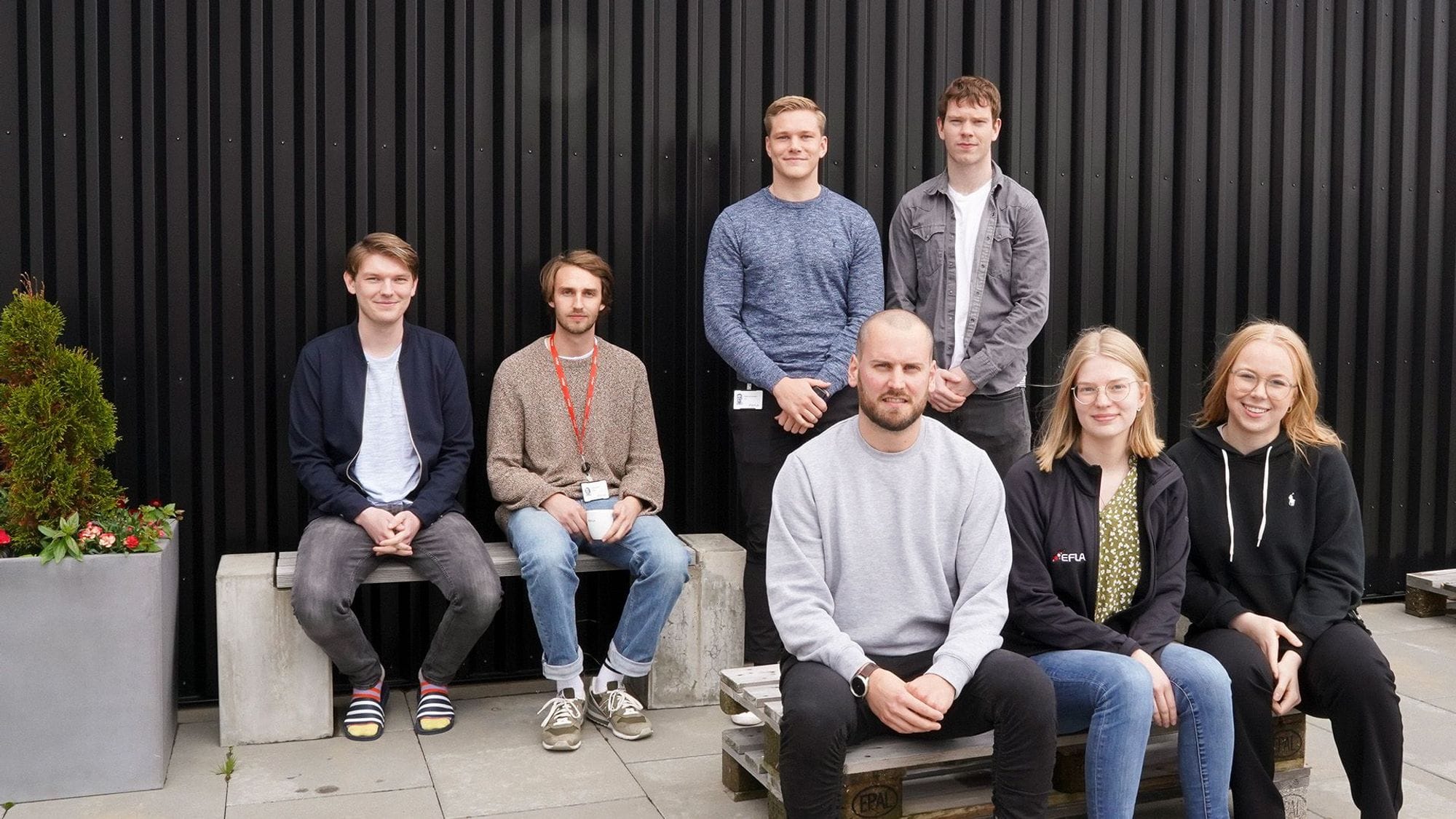 Seven individuals posing for a photo, against corrugated black wall backdrop
