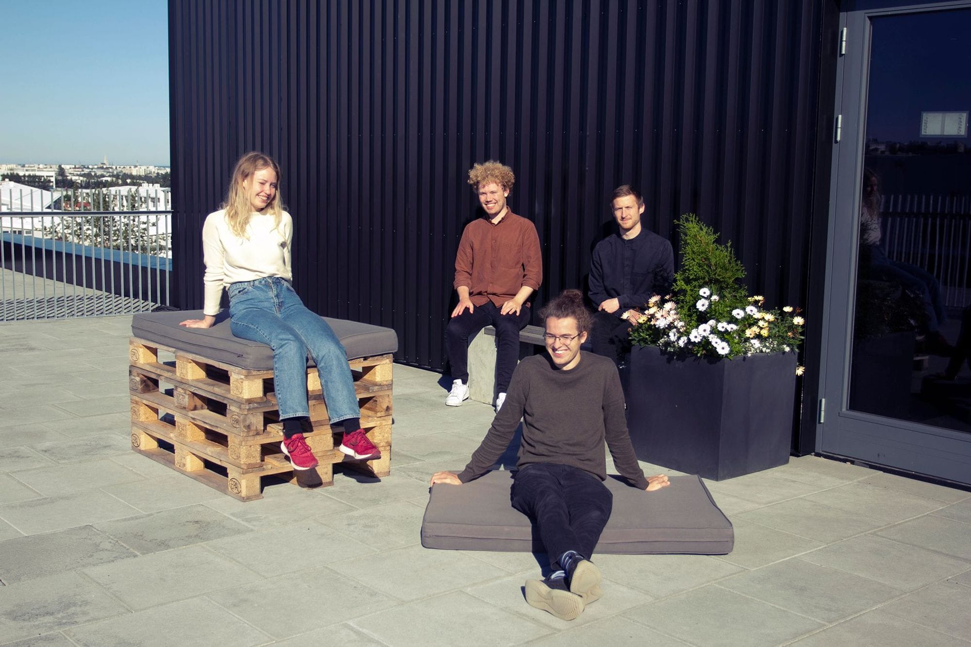 Four individuals relaxing on a sunny rooftop with seating made from pallets and cushions