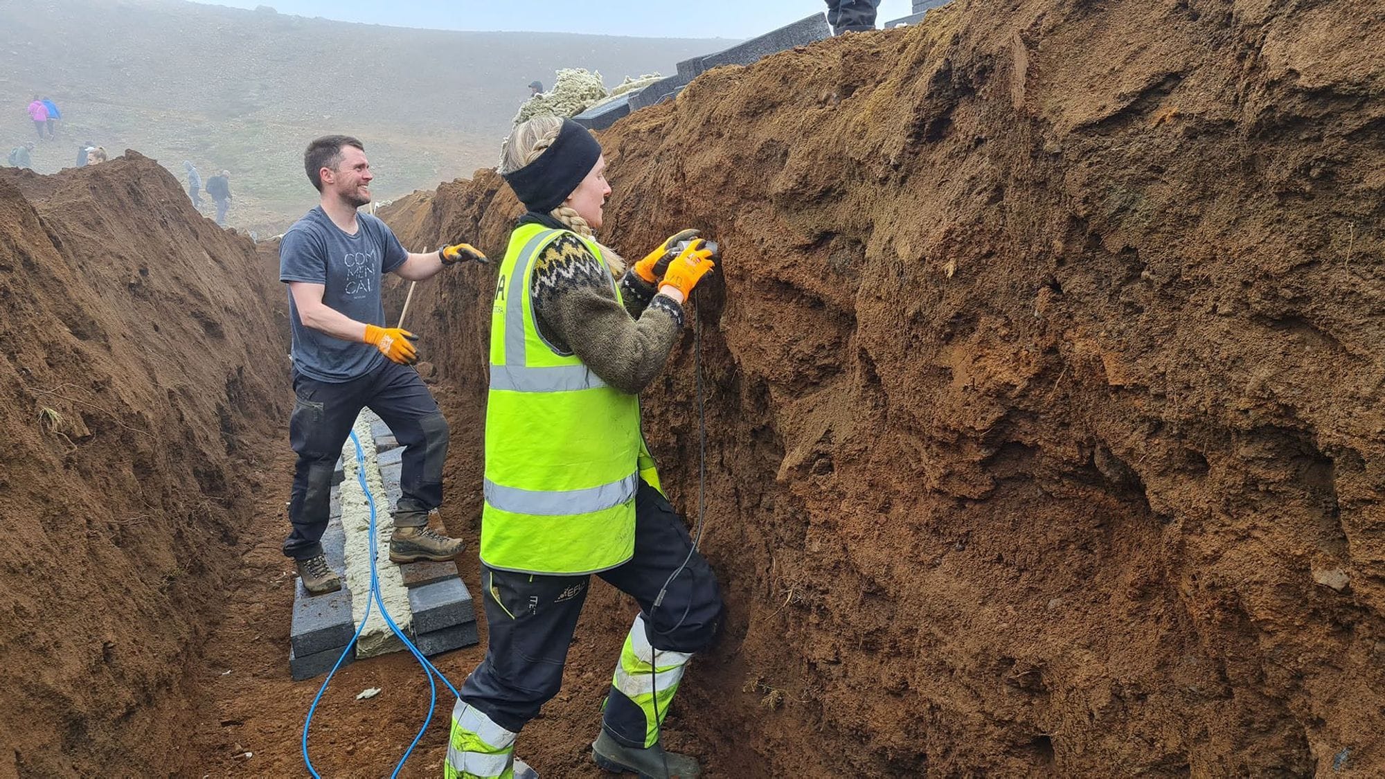 Two individuals in high visibility vest, working with tools inside a deep trench 