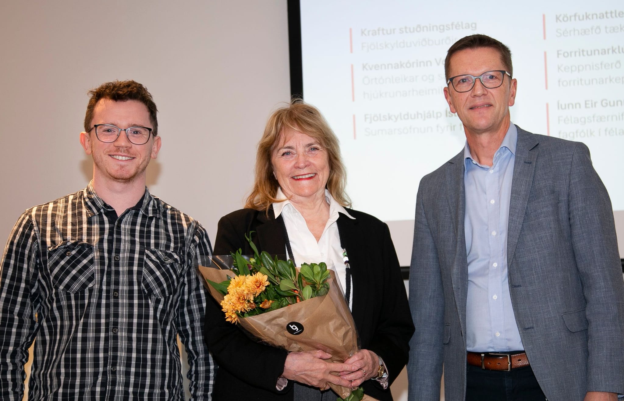 Three individuals standing in front of a presentation screen, with one person holding a bouquet of flowers