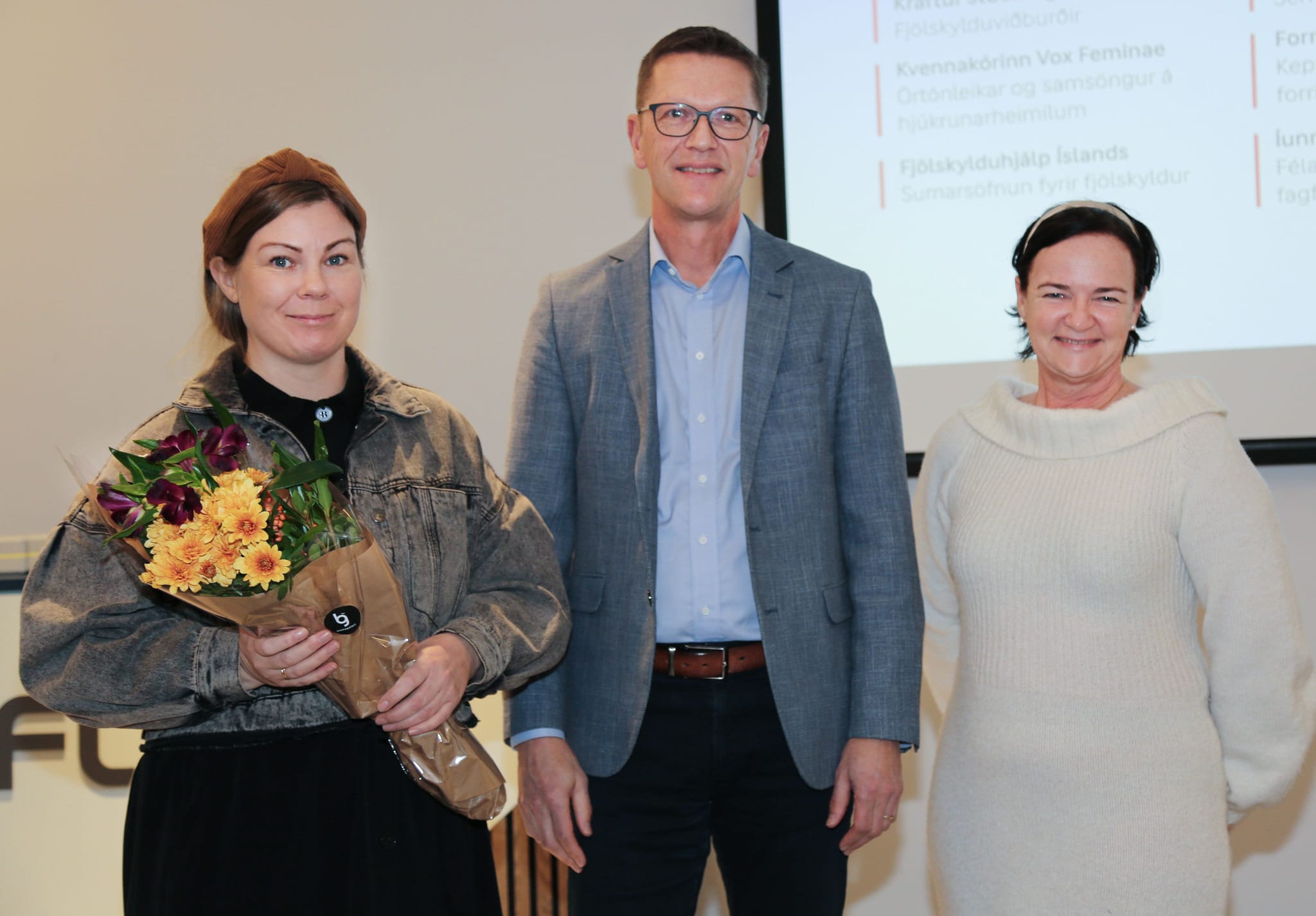 Three individuals standing in front of a presentation screen, with one person holding a bouquet of flowers