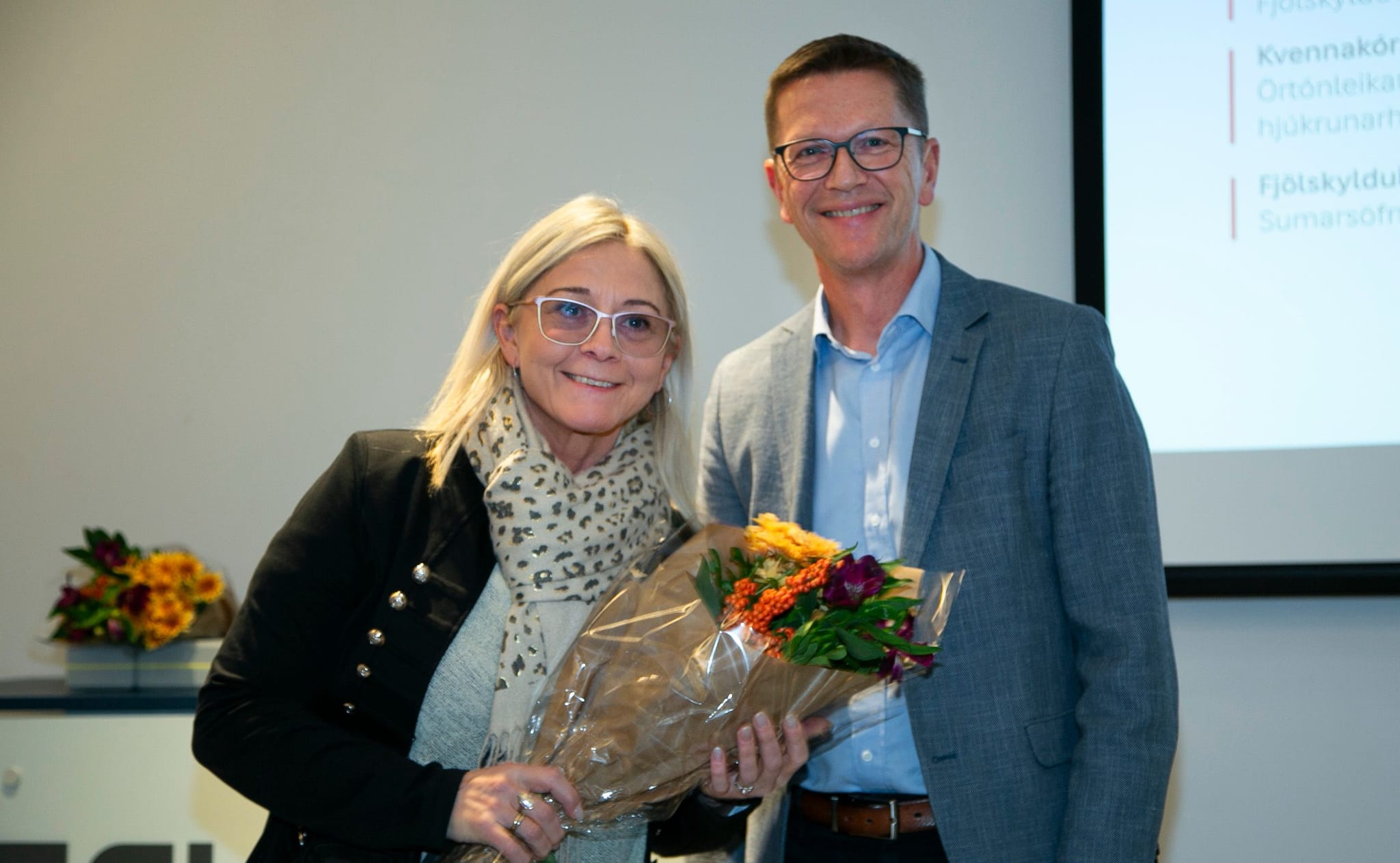 Two individuals standing in front of a presentation screen, with one person holding a bouquet of flowers