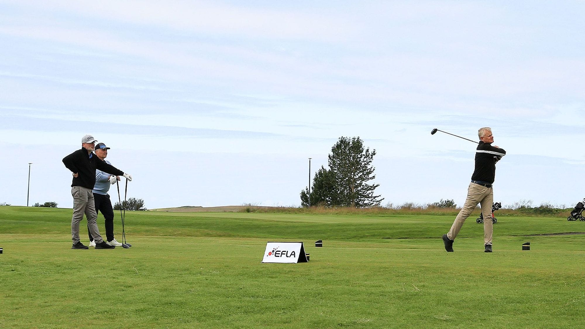Three men on a golf course, two of them are posing with their clubs and the third one in mid-swing