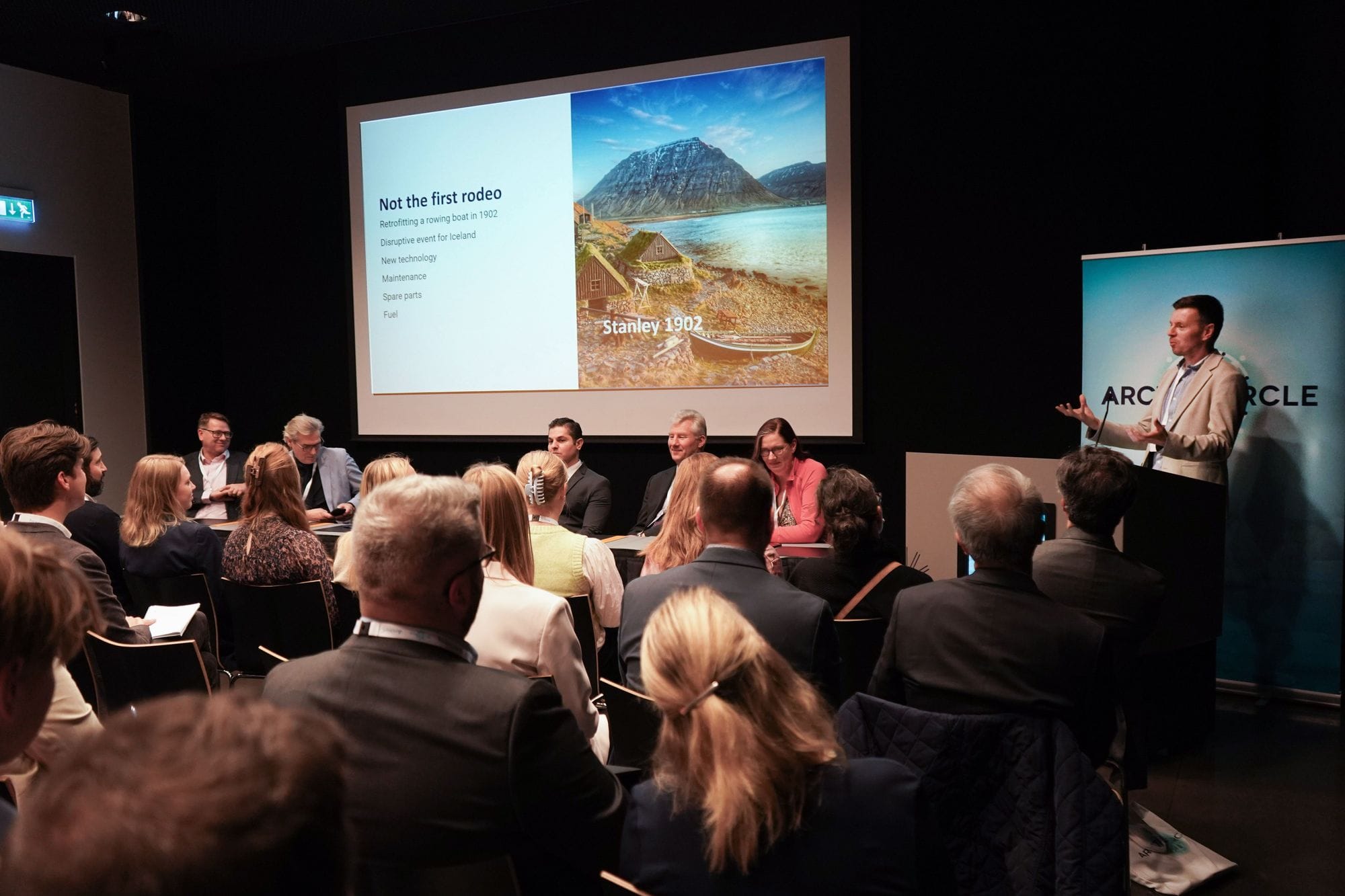 An audience facing a panel of speakers and a presenter standing next to a screen displaying a PowerPoint 