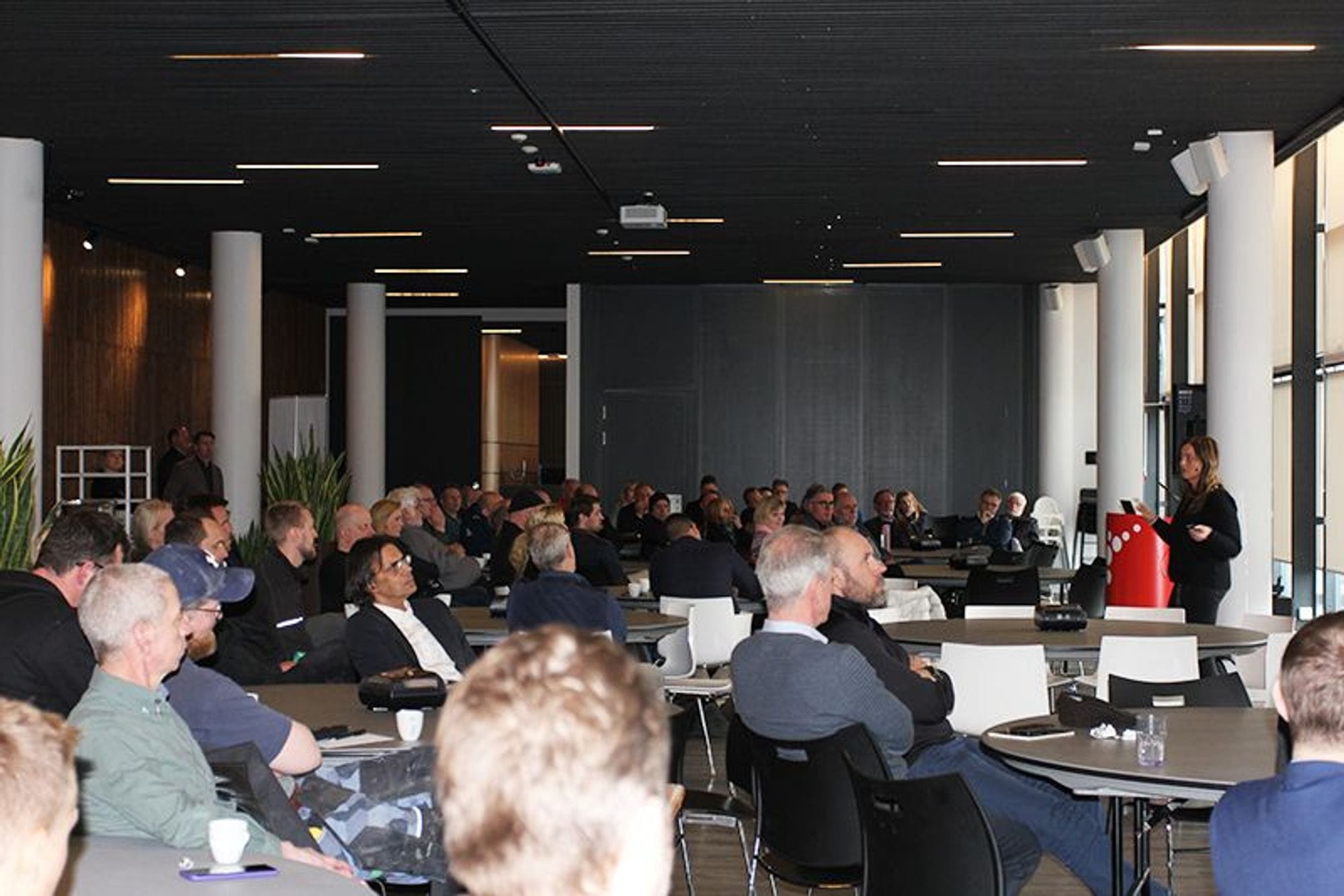 A large group of people sitting in a conference room with speaker visible in the distance