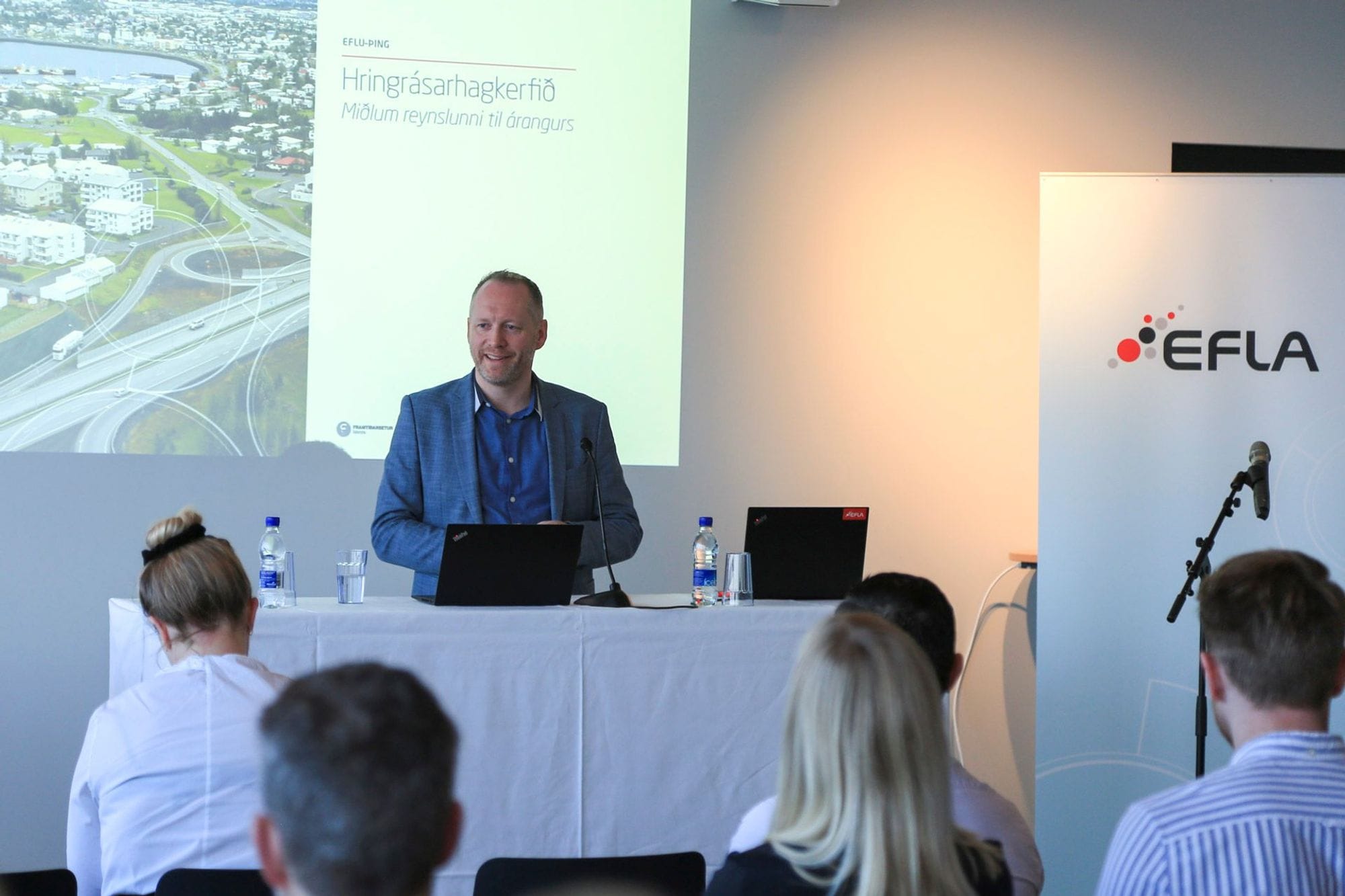 A man giving presentation to an audience with  two laptops on the table and a big screen in the background 