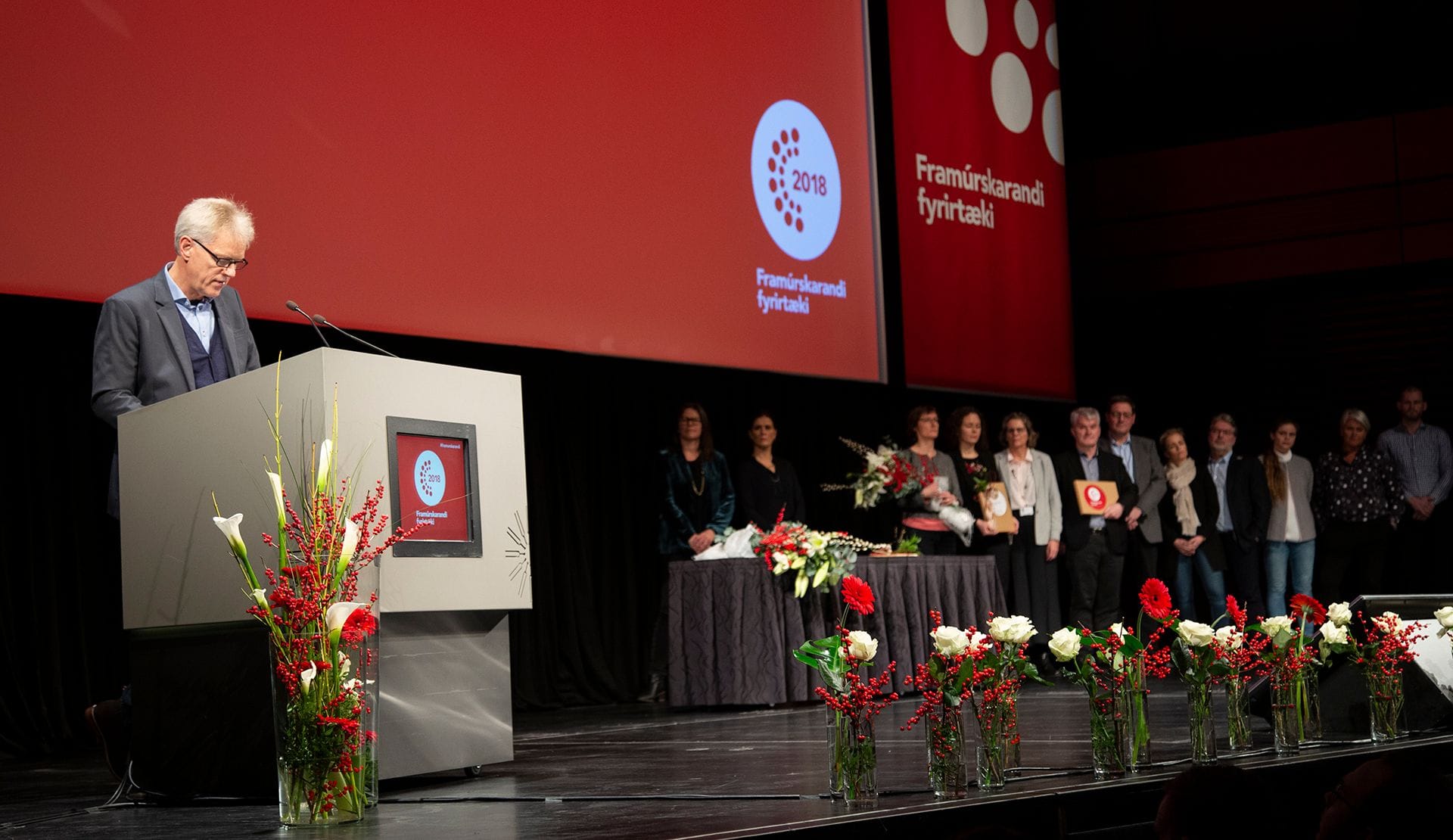 A man giving speech on a stage with a group of people standing on the side, holding bouquet and certificate