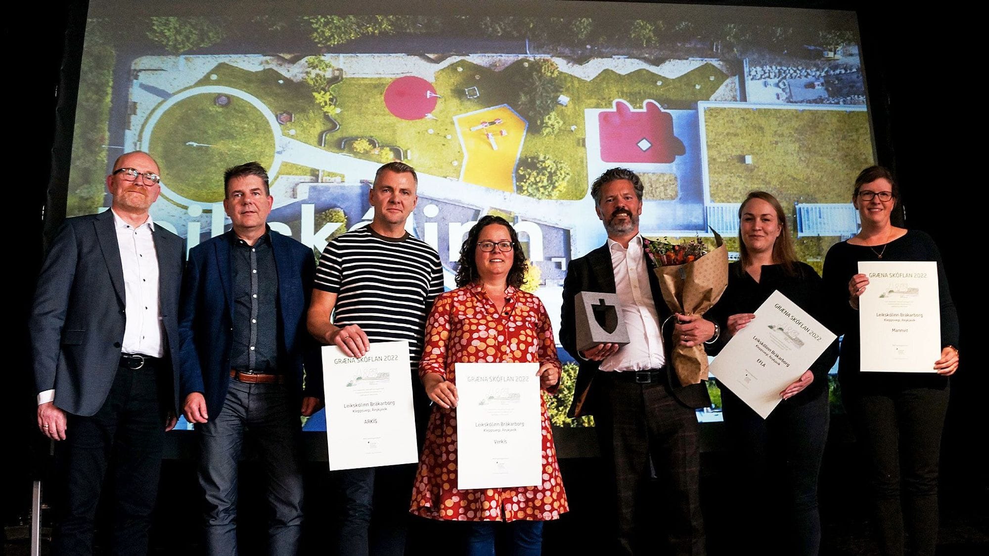 Six individuals holding certificates and flower bouquet with a screen in the background displaying a colorful layouts
