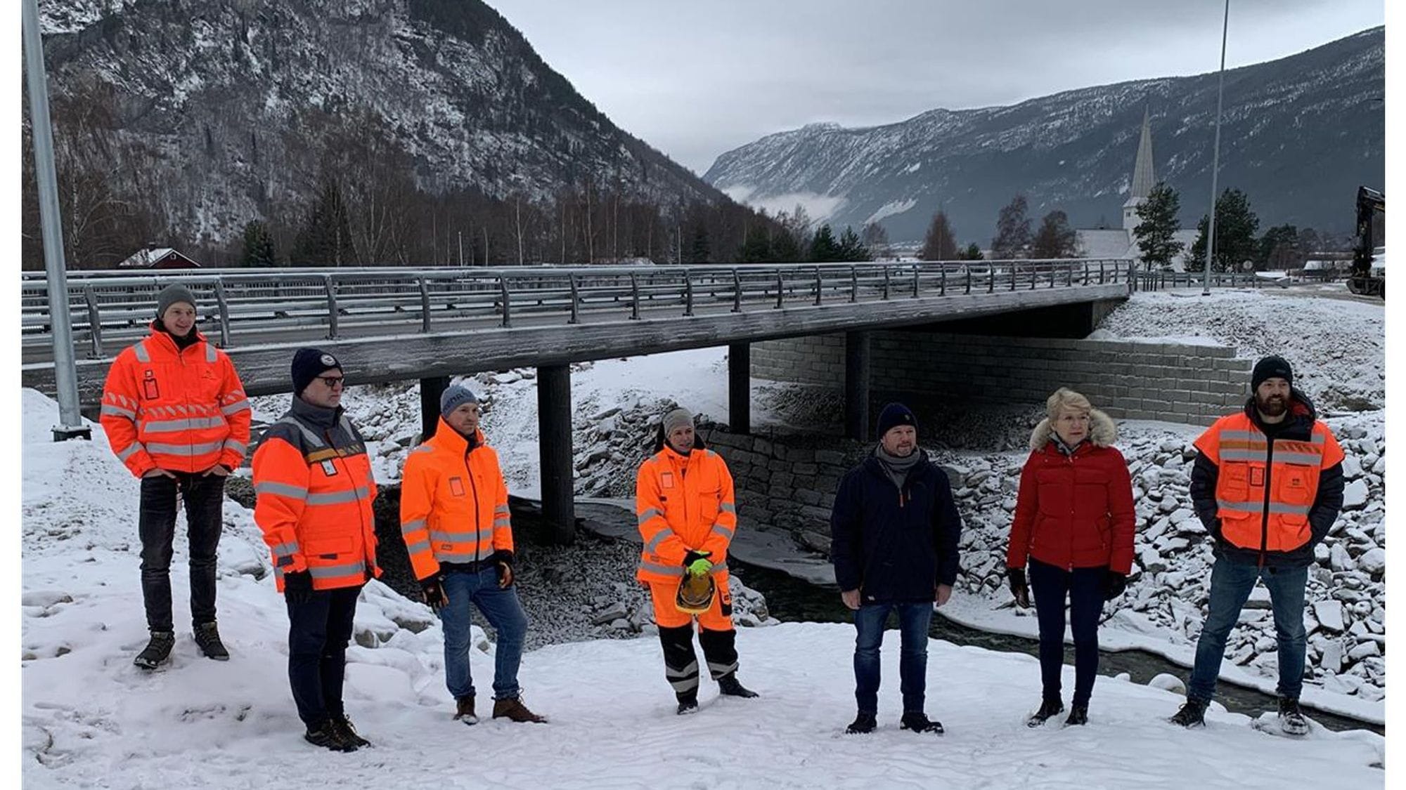 A group of people, some wearing high visibility orange jacket, standing in a snow with a mountain background