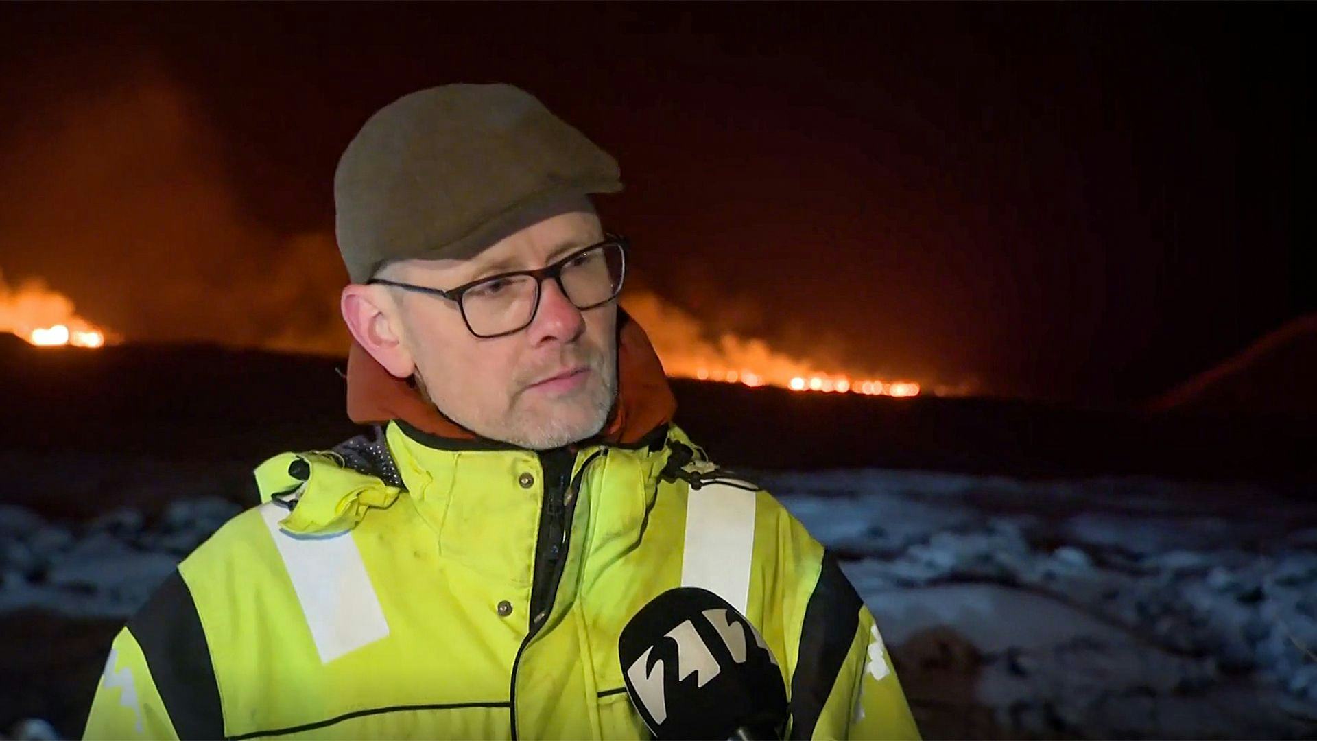 A man in high visibility jacket in the foreground with a dramatic background of what appears to be a fire or blaze in the night