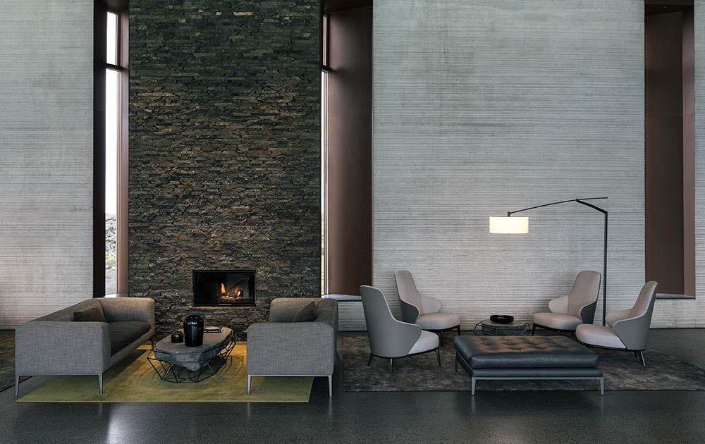 An image of a hotel lobby or similar setting which includes grey couches, coffee table, floor lamp and a fire place