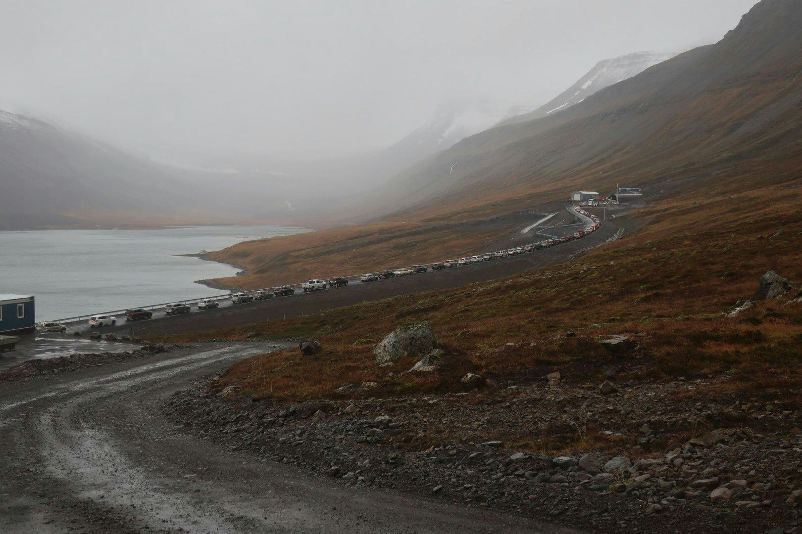A line of cars on a road besides a misty mountain and a lake