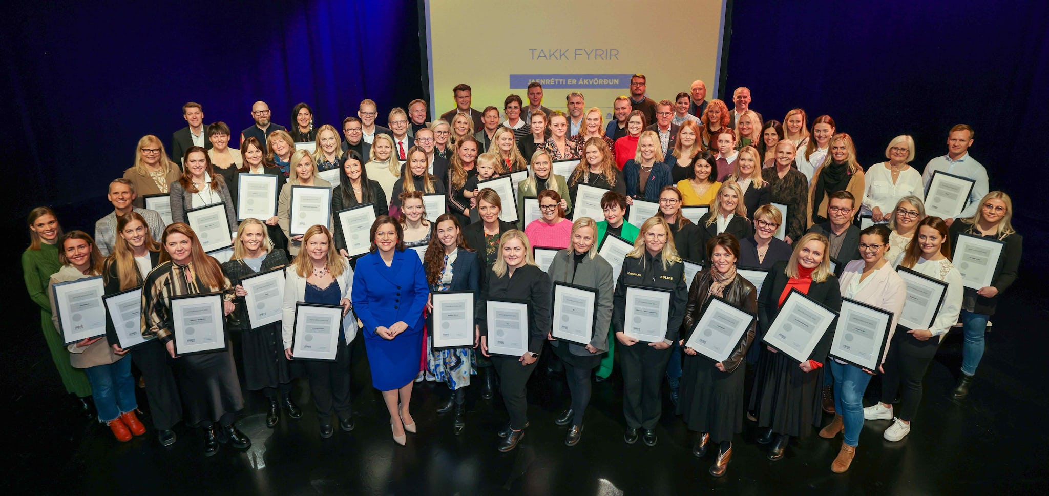 A large group of people posing together, many holding certificate with a yellow background that reads "TAKK FYRIR"