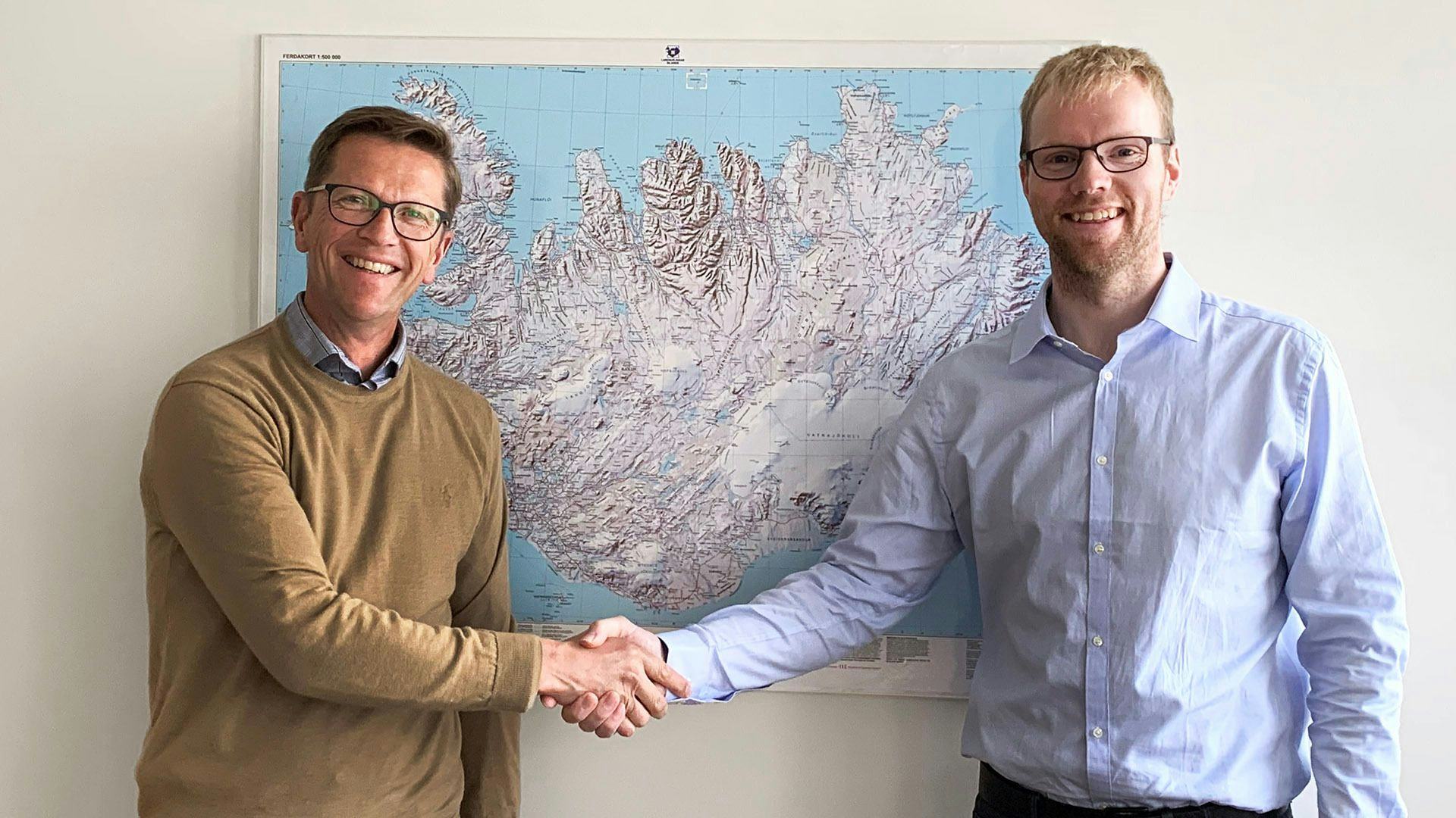 Two smiling men shaking hands in front of a large wall map
