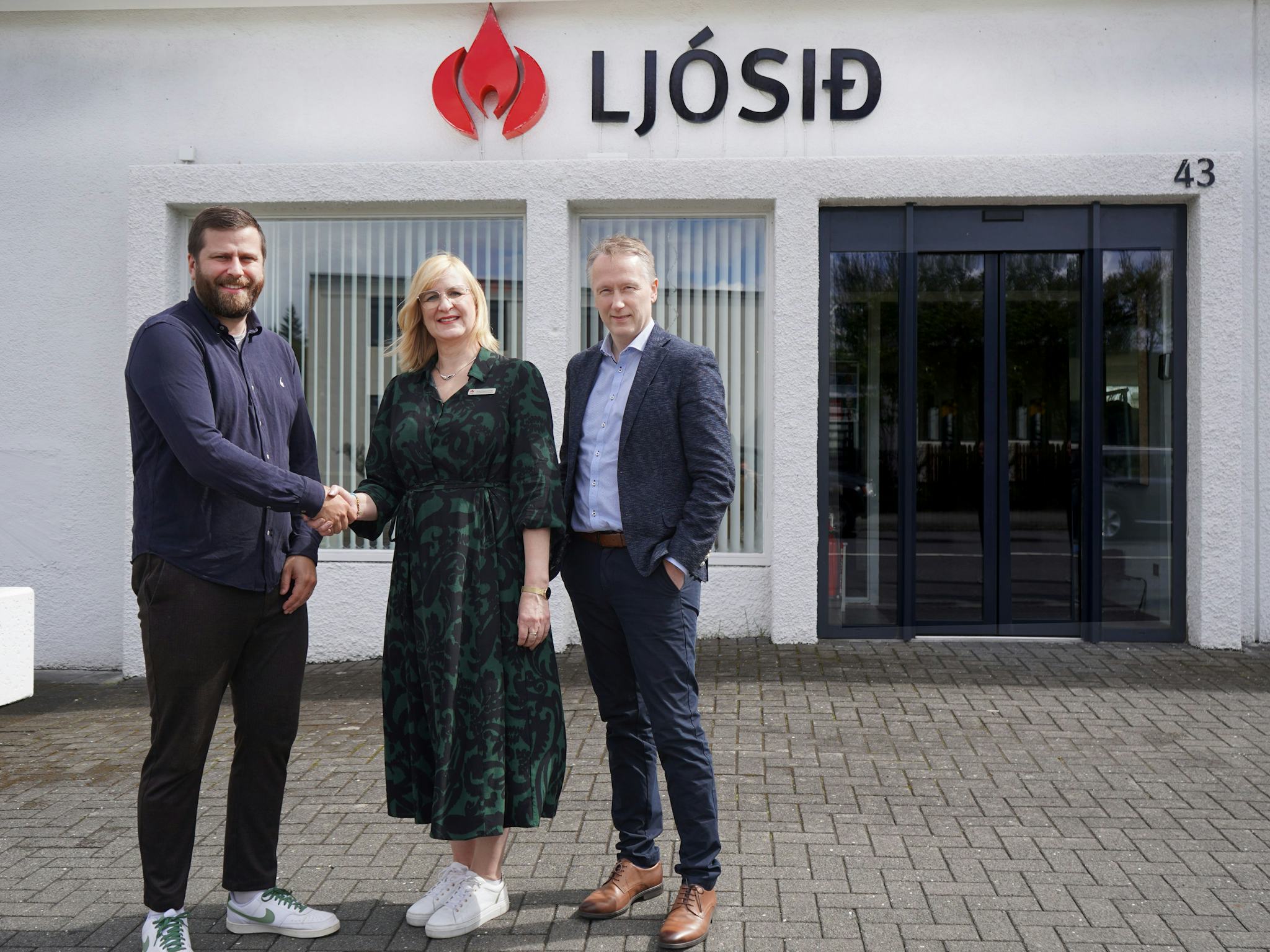 Three individuals standing together and shaking hands in front of a building with the sign "LJOSID"