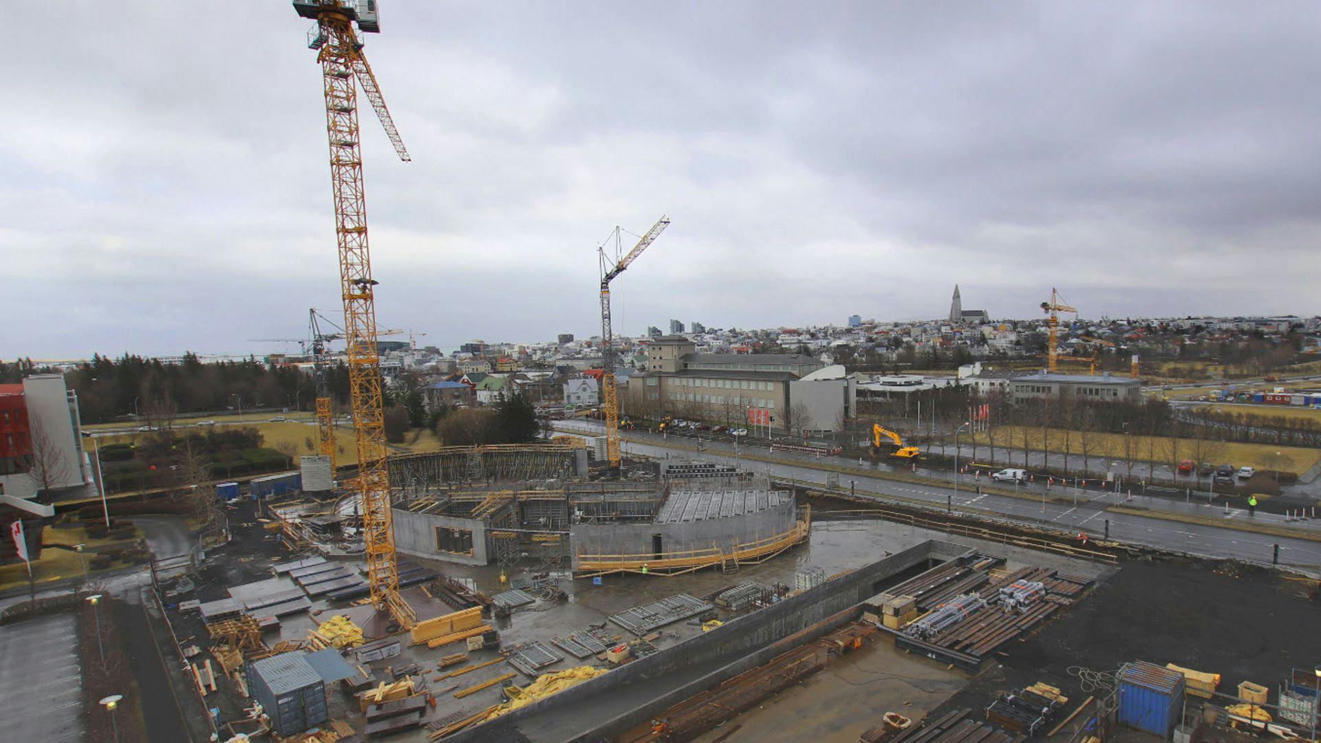An urban construction site with huge yellow cranes and a cityscape in the distance under overcast skies