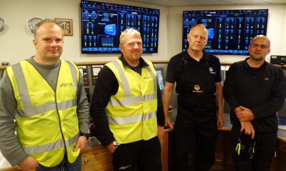 The photo captures four men, two men in high visibility vest, standing in front of a control panel with numerous digital display