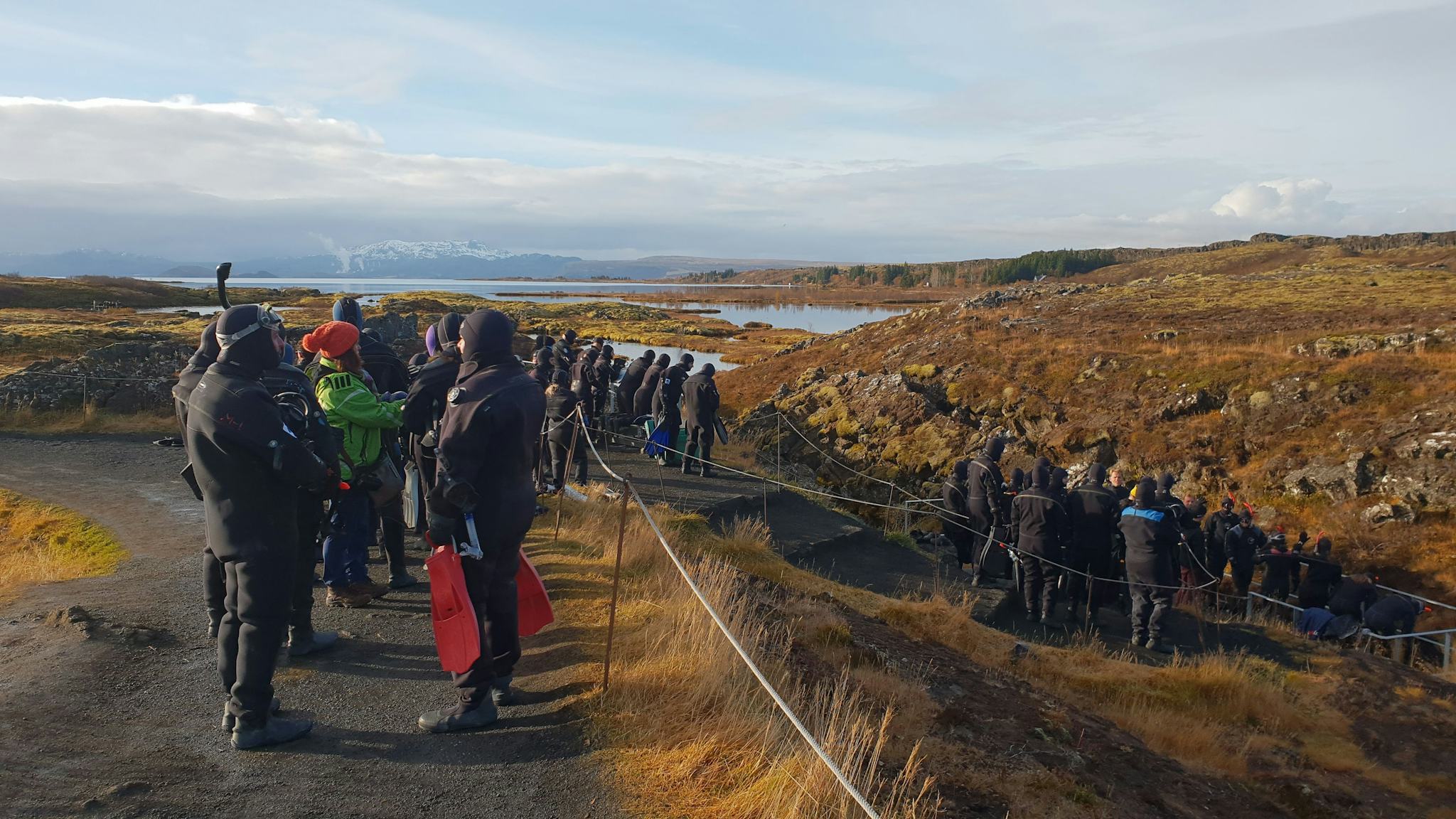 A crowd dressed in warm clothes, queuing or walking along a pathway. The scenery includes a lake and landscape of grasses.