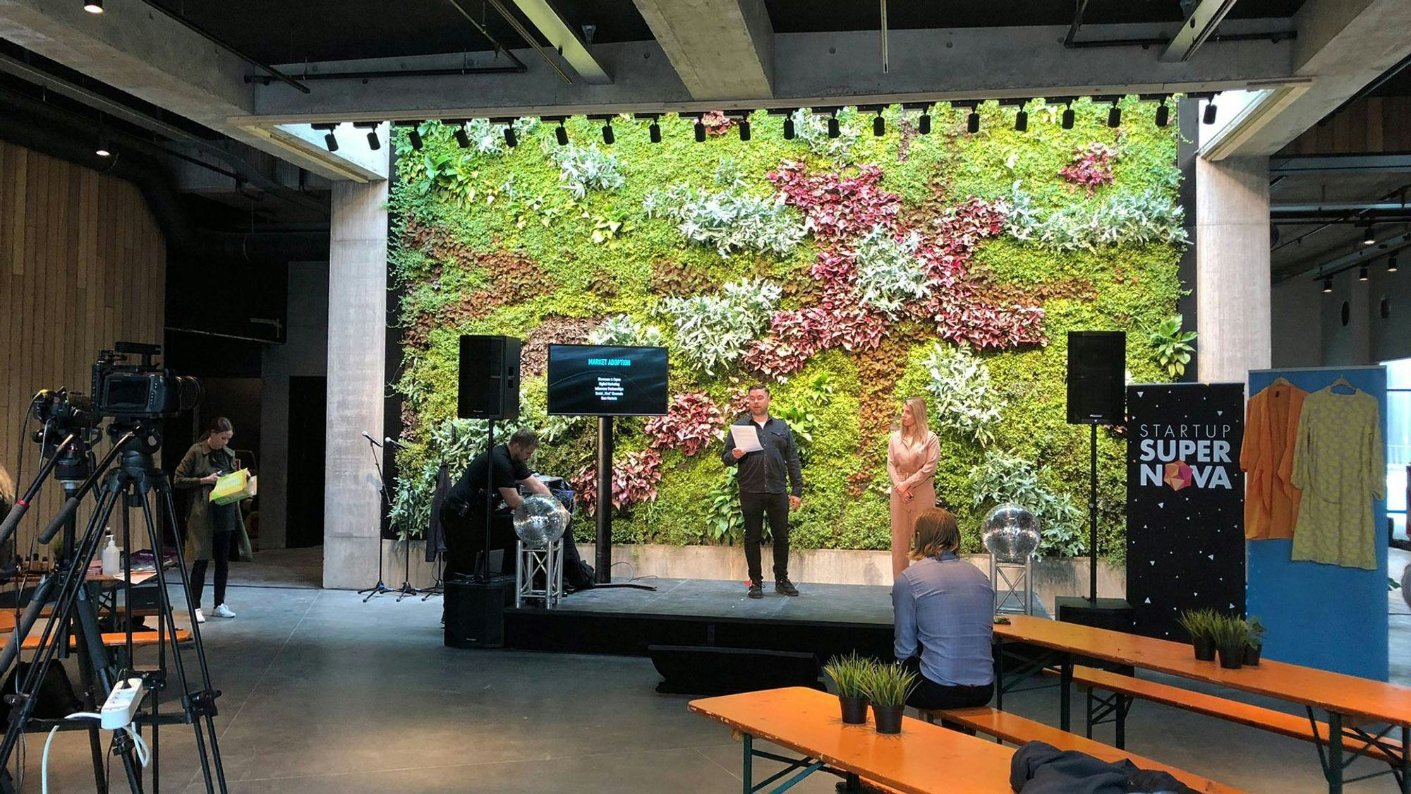 An indoor event with a man speaking next to a woman on a stage and a vertical garden wall as a backdrop