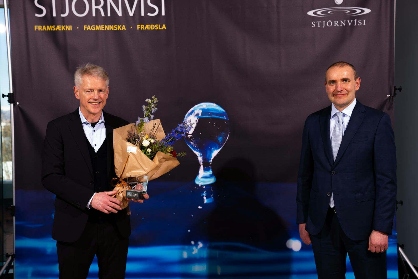 Two men in suits, one holding a bouquet of flowers, standing in front of a backdrop with the word "Stjórnvísi" and other texts