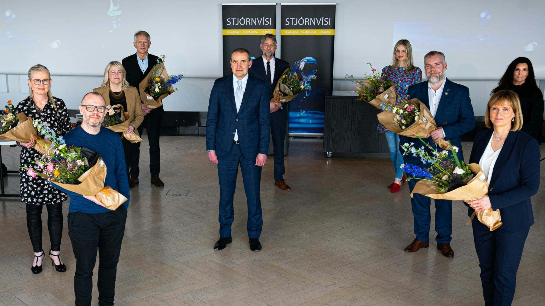 A group of ten individuals, each holding a bouquet of flowers, standing in a room with a backdrop that reads "Stjórnvísi"
