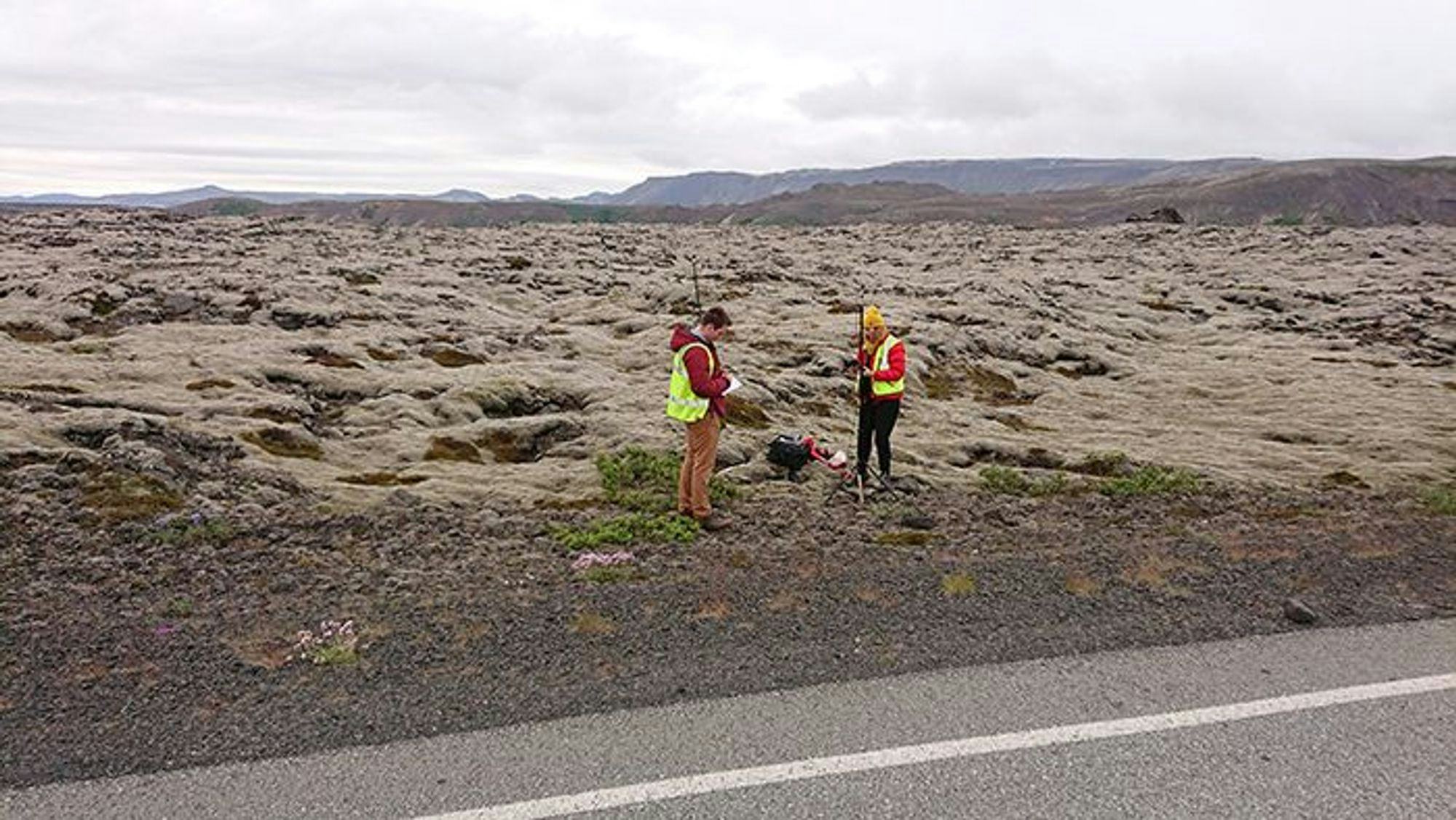 Two individuals in visibility vest by a roadside in a barren landscape