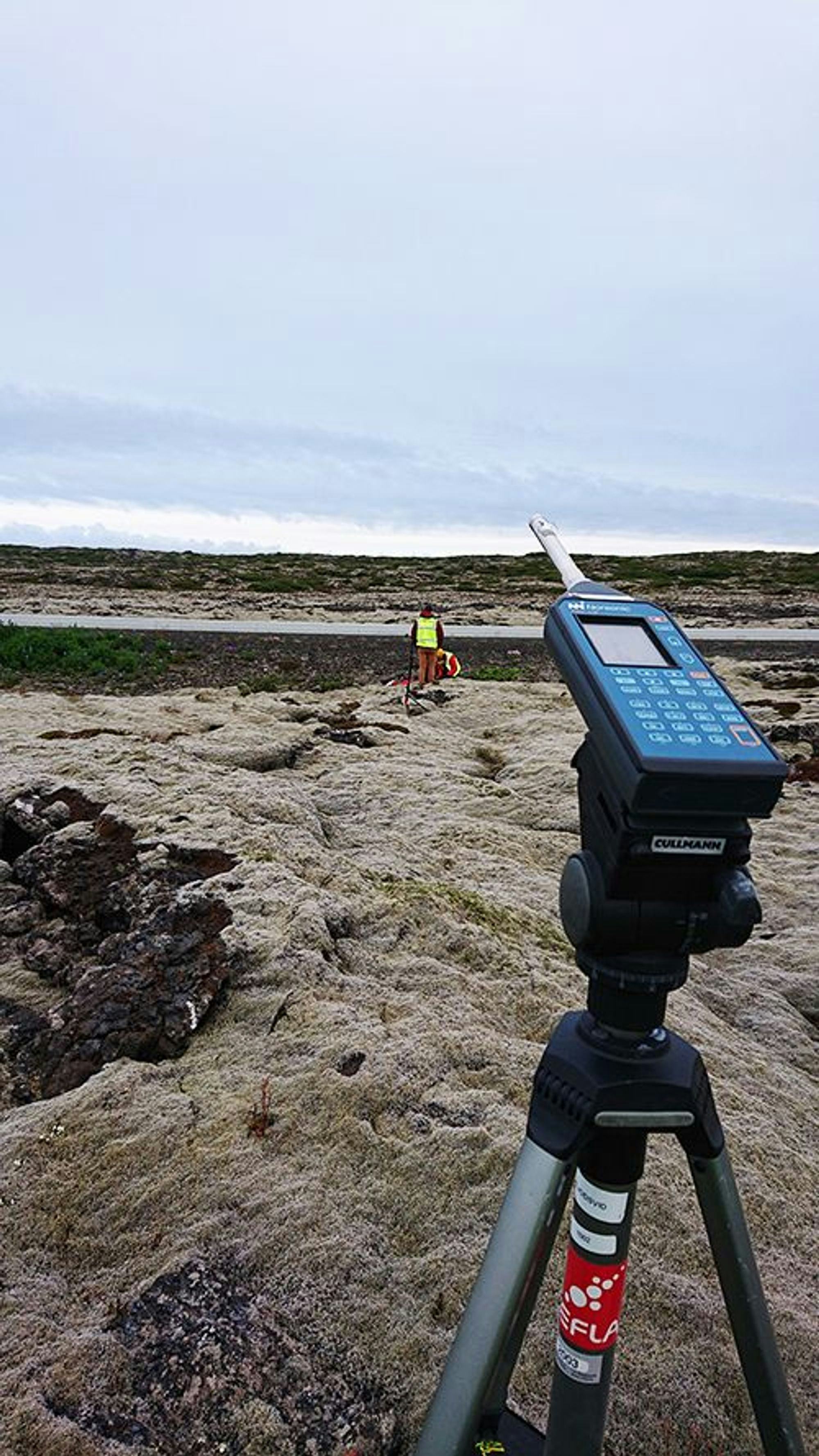 An image shows a device with buttons and a person working on a rocky terrain with sparse grass