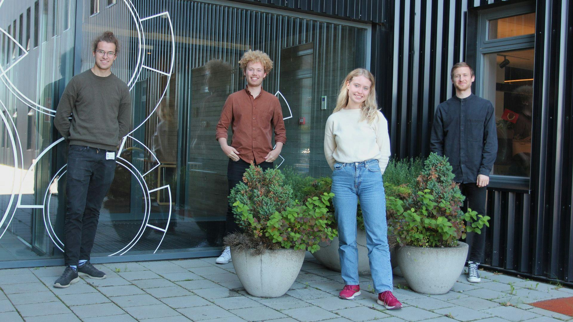 Four individuals casually standing outdoors next to large potted plants and decorative metal work