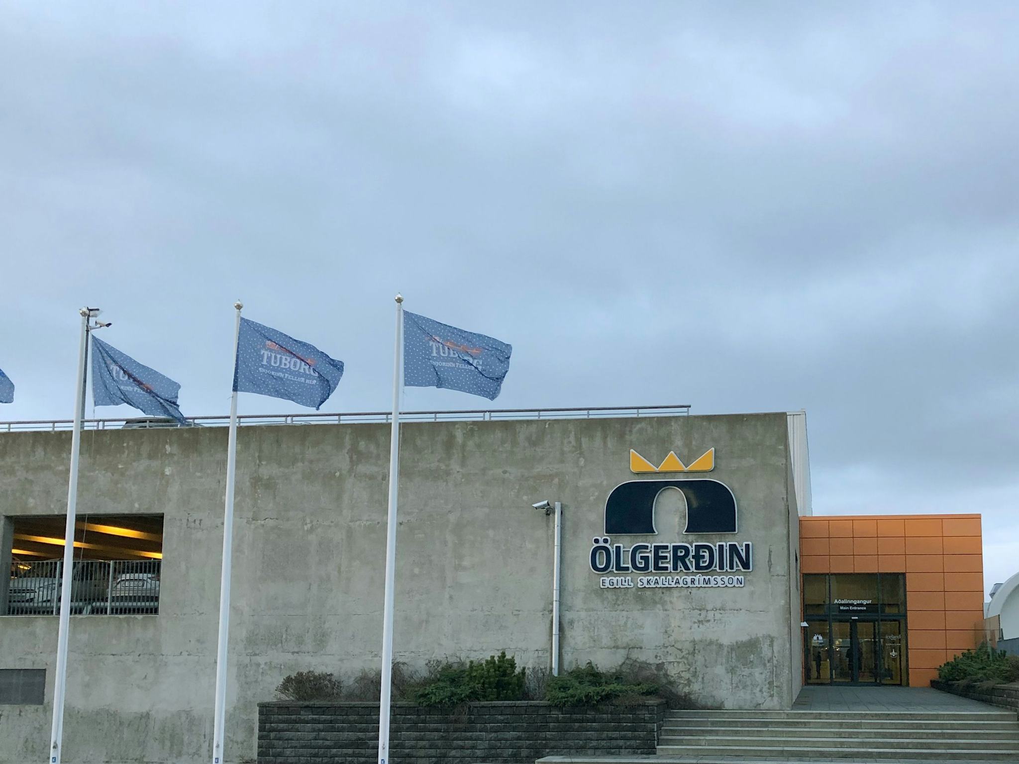 The image displays the front of a building with "Olgerdin" logo and three flags fluttering in the wind