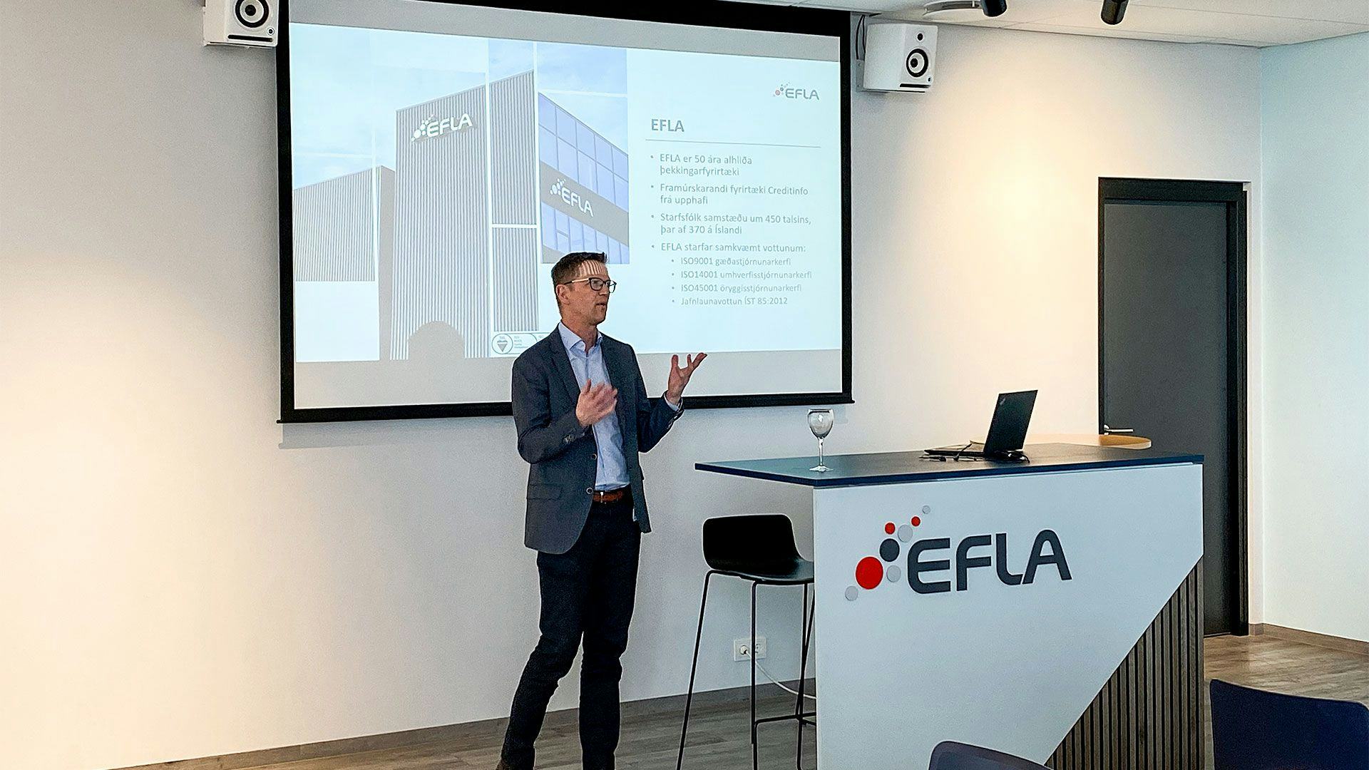 A man speaking at a podium labelled "EFLA" with a screen showing information about EFLA in the background
