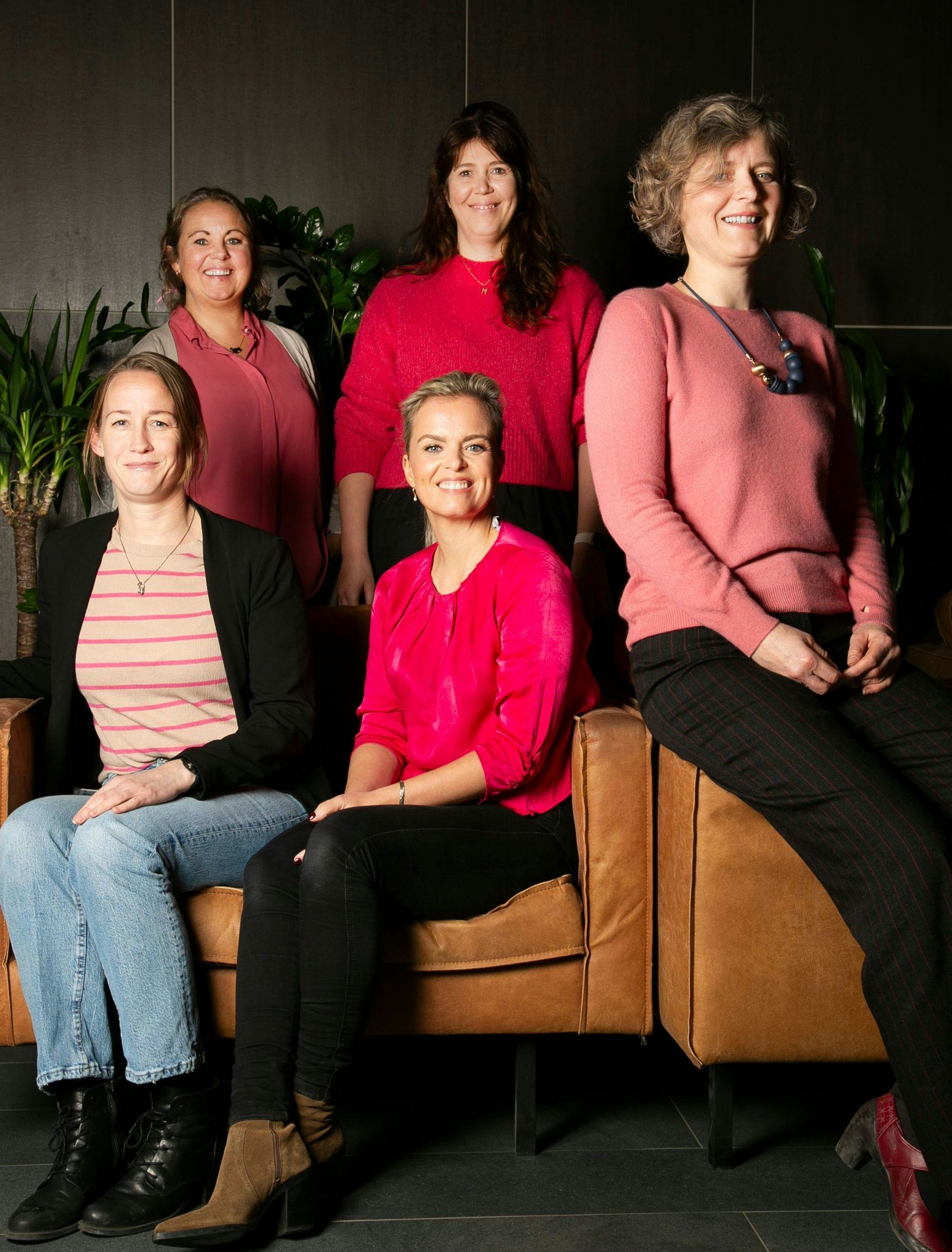 A group of five women, two standing and three seated on a leather couch, all set against a dark interior background with green foliage